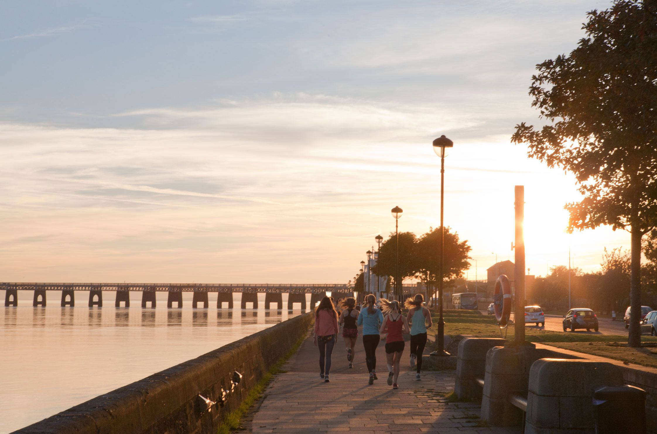 A group of runners jogs along a riverside path at sunset, with the warm golden light reflecting off the water and a distant bridge extending across the calm river.