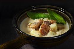 A bowl of cullen skink garnished with cooked meat, potato chunks, and fresh green leaves. The dish is served in a rustic dark bowl against a dark, moody background.
