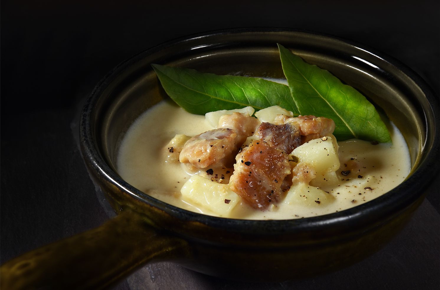 A bowl of cullen skink garnished with cooked meat, potato chunks, and fresh green leaves. The dish is served in a rustic dark bowl against a dark, moody background.
