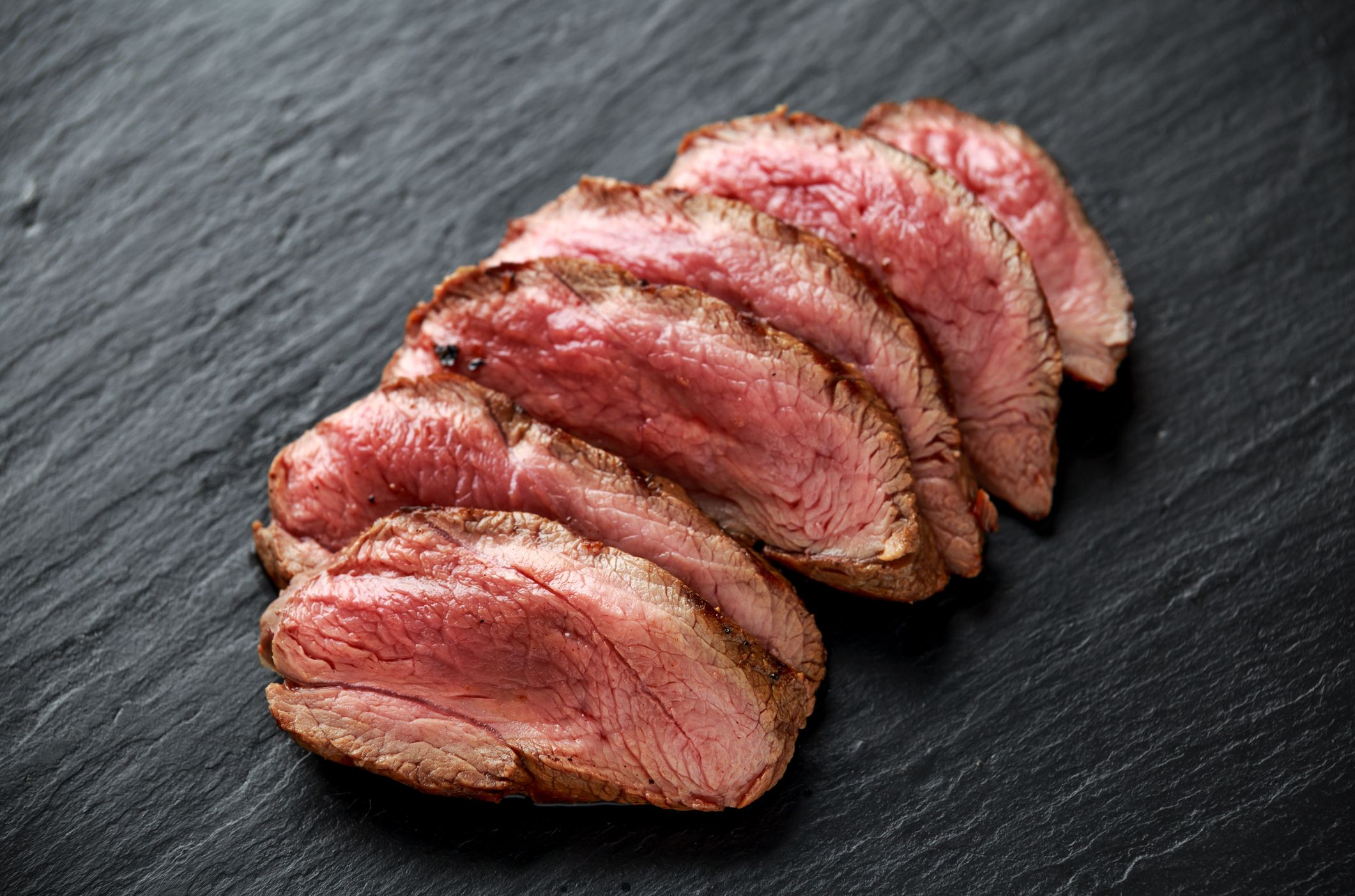 Slices of medium-rare roasted beef arranged on a dark slate surface, showcasing a juicy pink center and a seared brown crust.