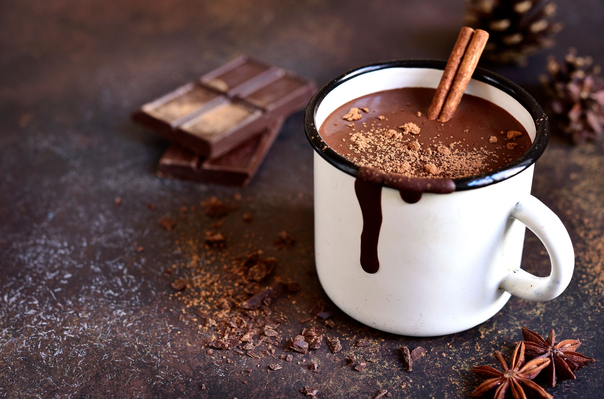 A white mug filled with rich hot chocolate, topped with cinnamon sticks and sprinkled cocoa powder. Melted chocolate drips down the side, with chocolate bars and spices nearby.