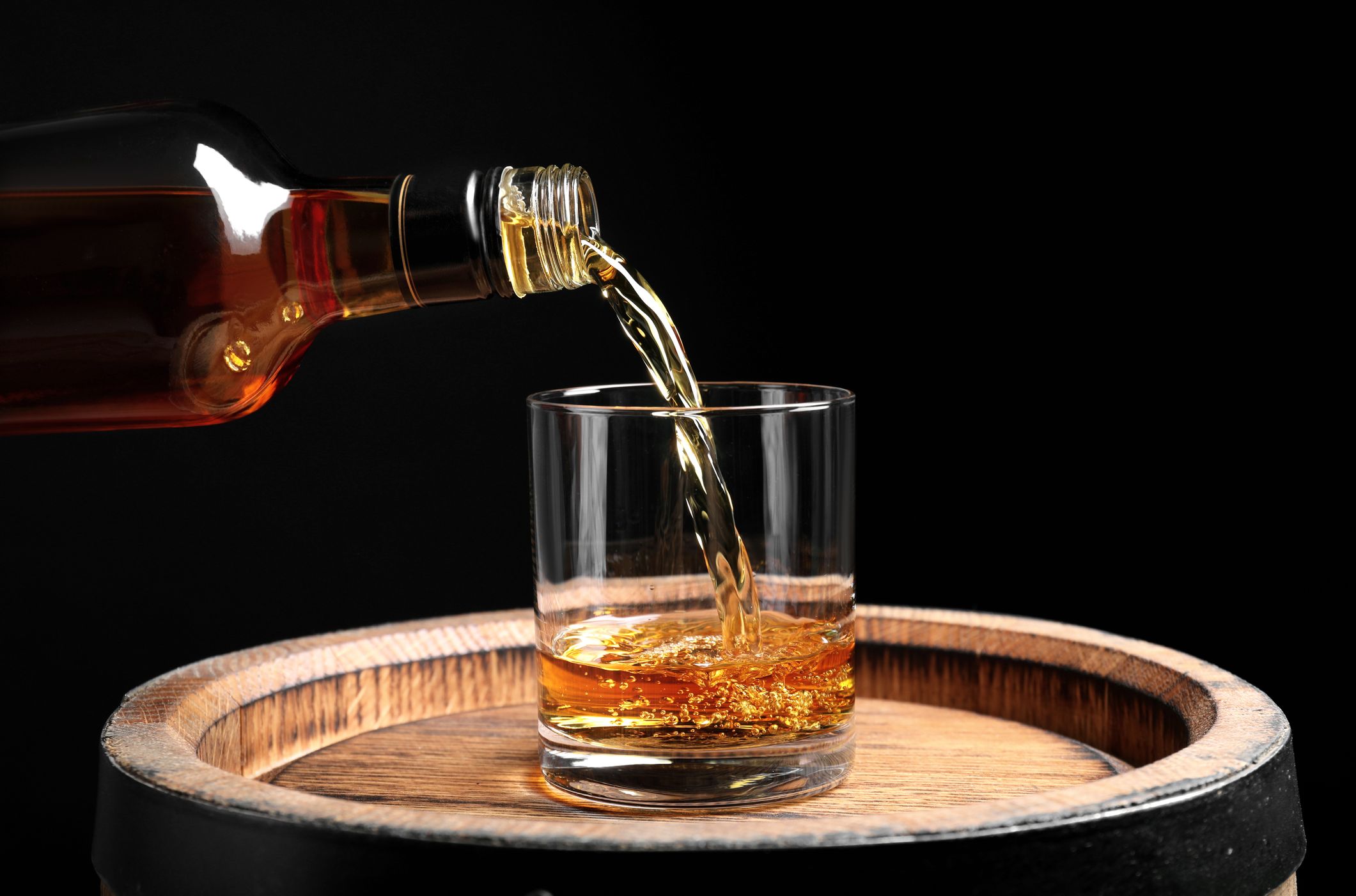 A glass of whiskey being poured from a bottle, placed on a wooden barrel against a dark background. The golden liquid creates ripples as it fills the clear glass.