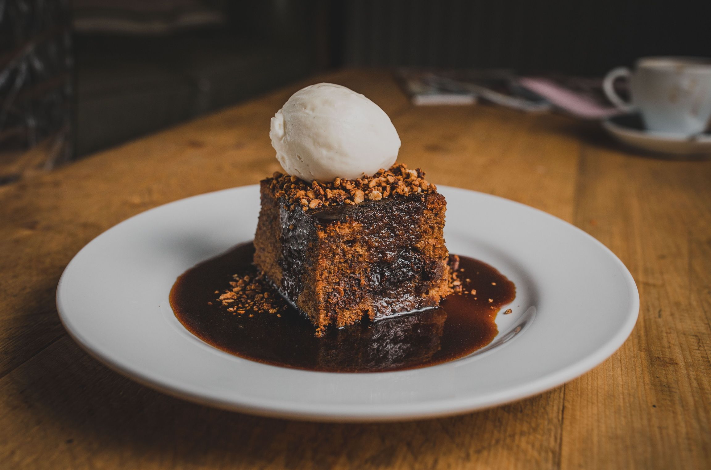 A rich, sticky toffee pudding topped with a scoop of vanilla ice cream, served on a white plate with caramel sauce drizzled around the dessert.