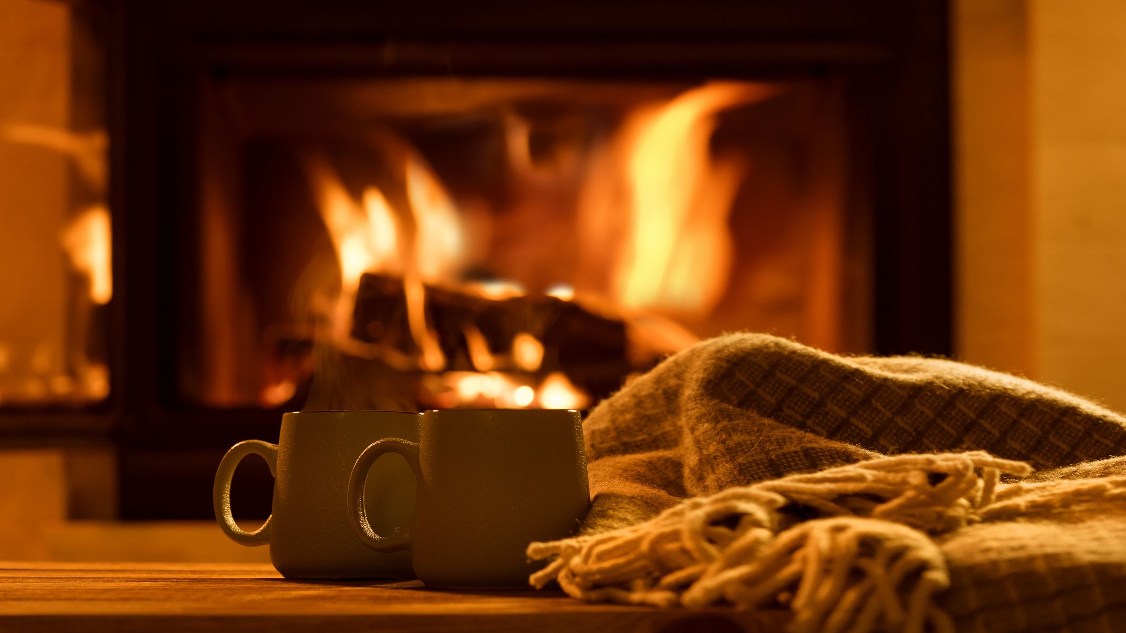 Two mugs sit on a wooden surface next to a cozy blanket in front of a crackling fireplace. The warm glow of the fire creates a comforting and inviting atmosphere.