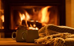 Two mugs sit on a wooden surface next to a cozy blanket in front of a crackling fireplace. The warm glow of the fire creates a comforting and inviting atmosphere.