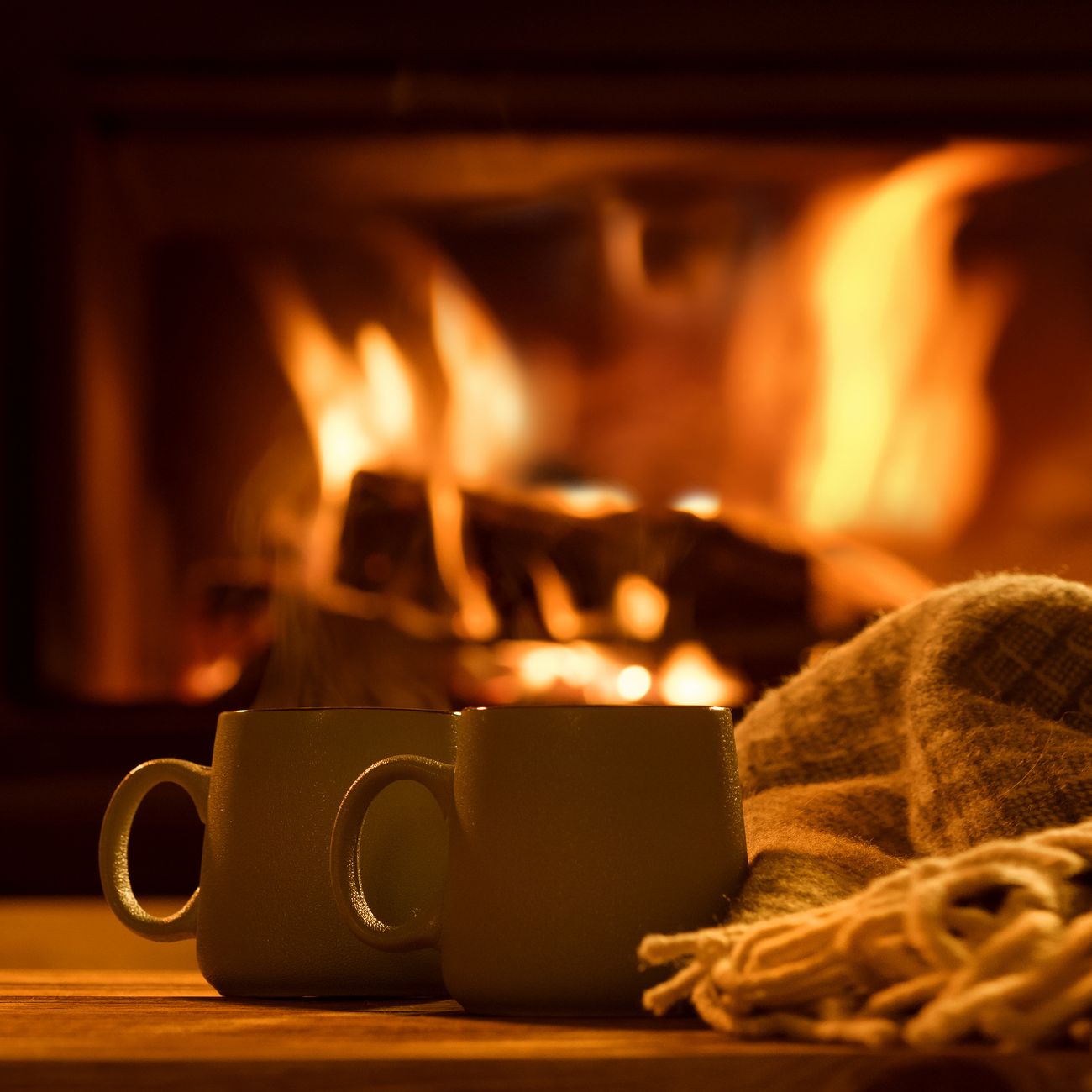 Two mugs sit on a wooden surface next to a cozy blanket in front of a crackling fireplace. The warm glow of the fire creates a comforting and inviting atmosphere.
