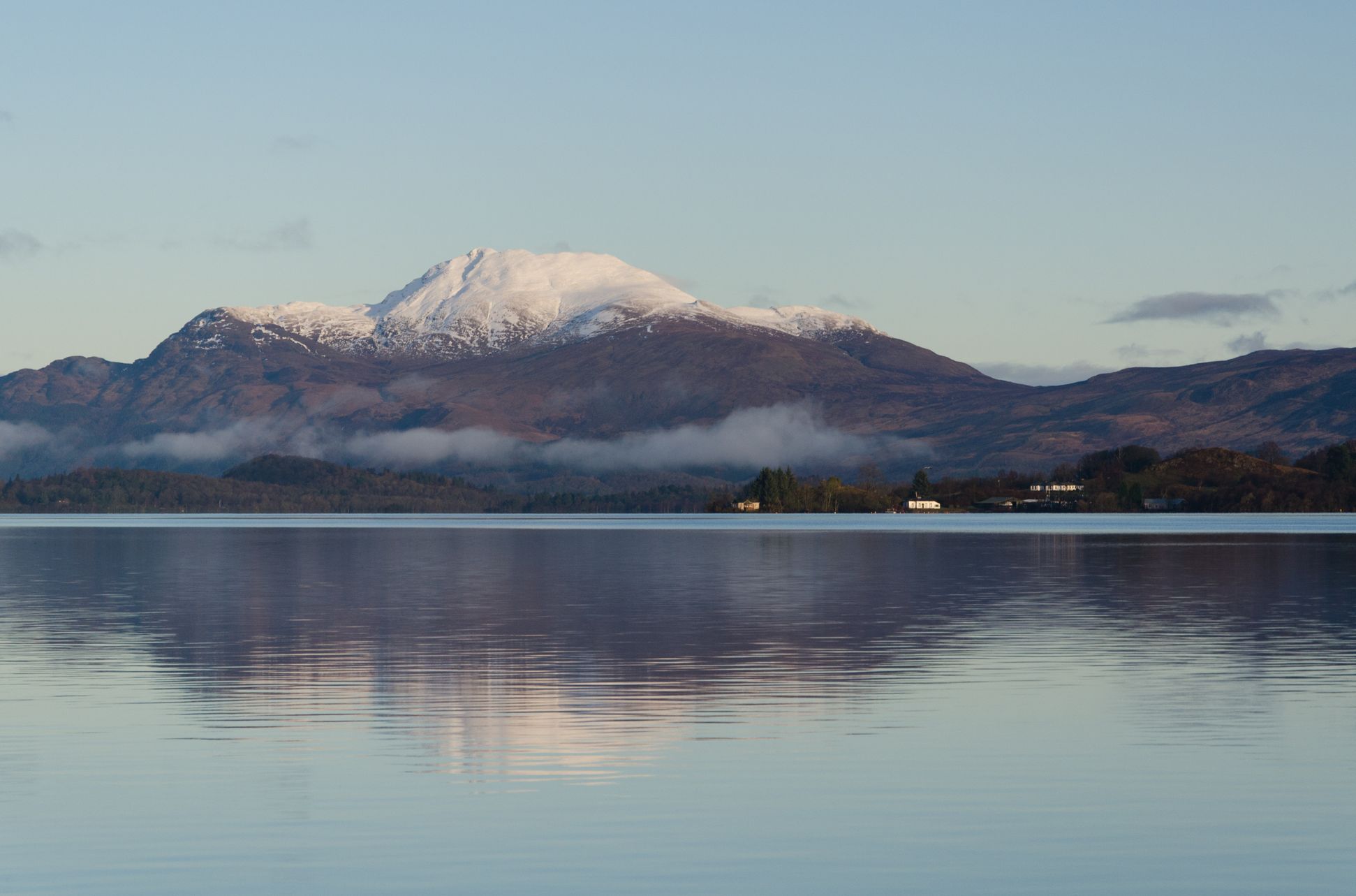 A calm lake reflects snow-capped mountains under a clear sky, with mist hovering over the forested hills in the distance, creating a peaceful, serene landscape.