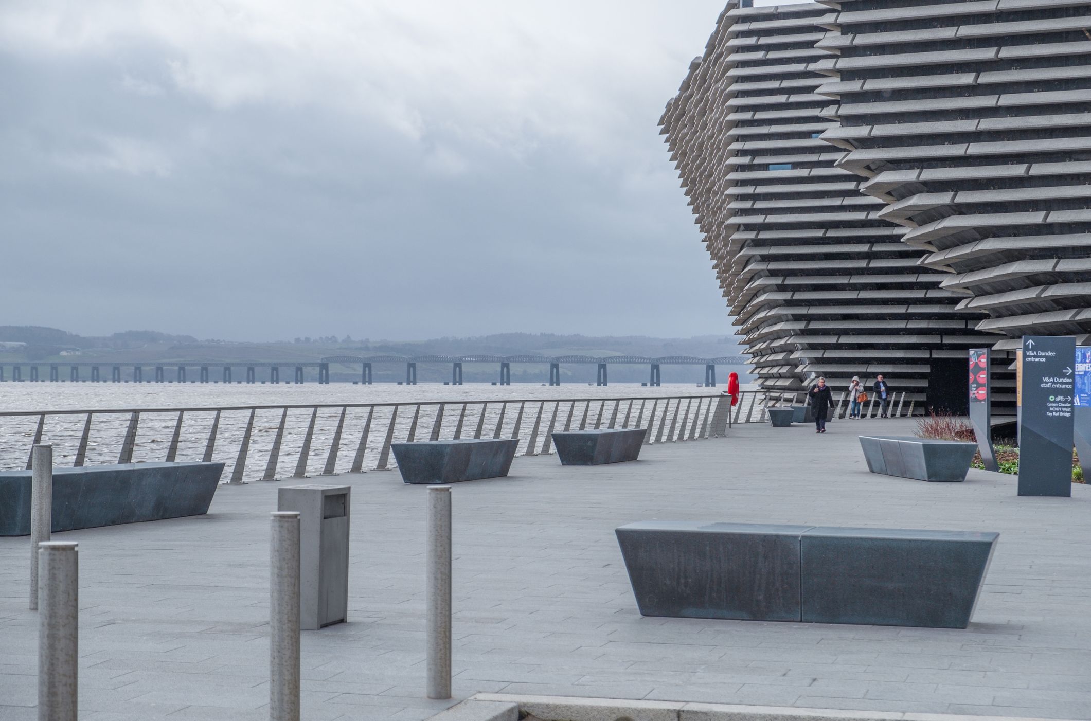 A modern architectural building with layered design stands beside a waterfront promenade, featuring benches, a railing, and a distant bridge under a cloudy sky.