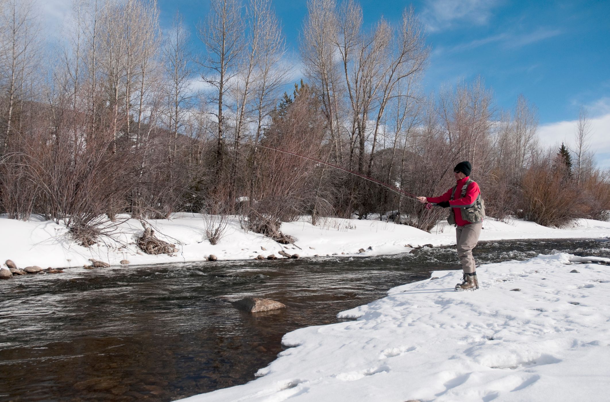 A person in a red jacket and winter gear fly-fishing on a snowy riverbank, with bare trees and a flowing river under a clear blue sky in the background.