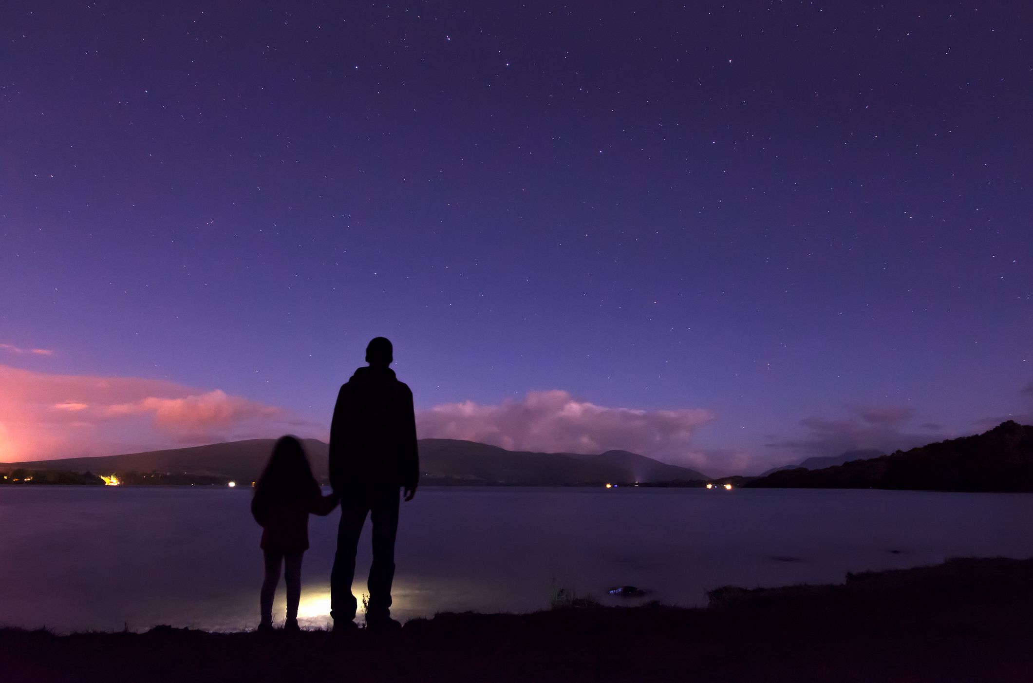 Silhouettes of an adult and child standing by a calm lake at night, gazing at a star-filled sky with distant hills and soft clouds faintly illuminated in the background.