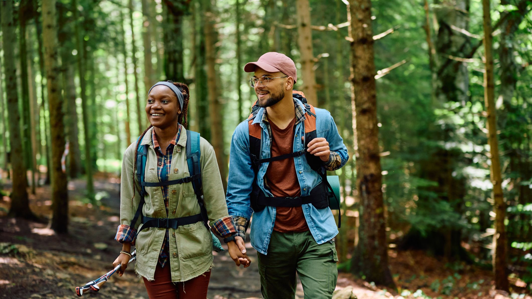 A smiling couple hiking through a lush forest, holding hands and wearing backpacks. The woman carries a trekking pole, and dappled sunlight filters through the trees.