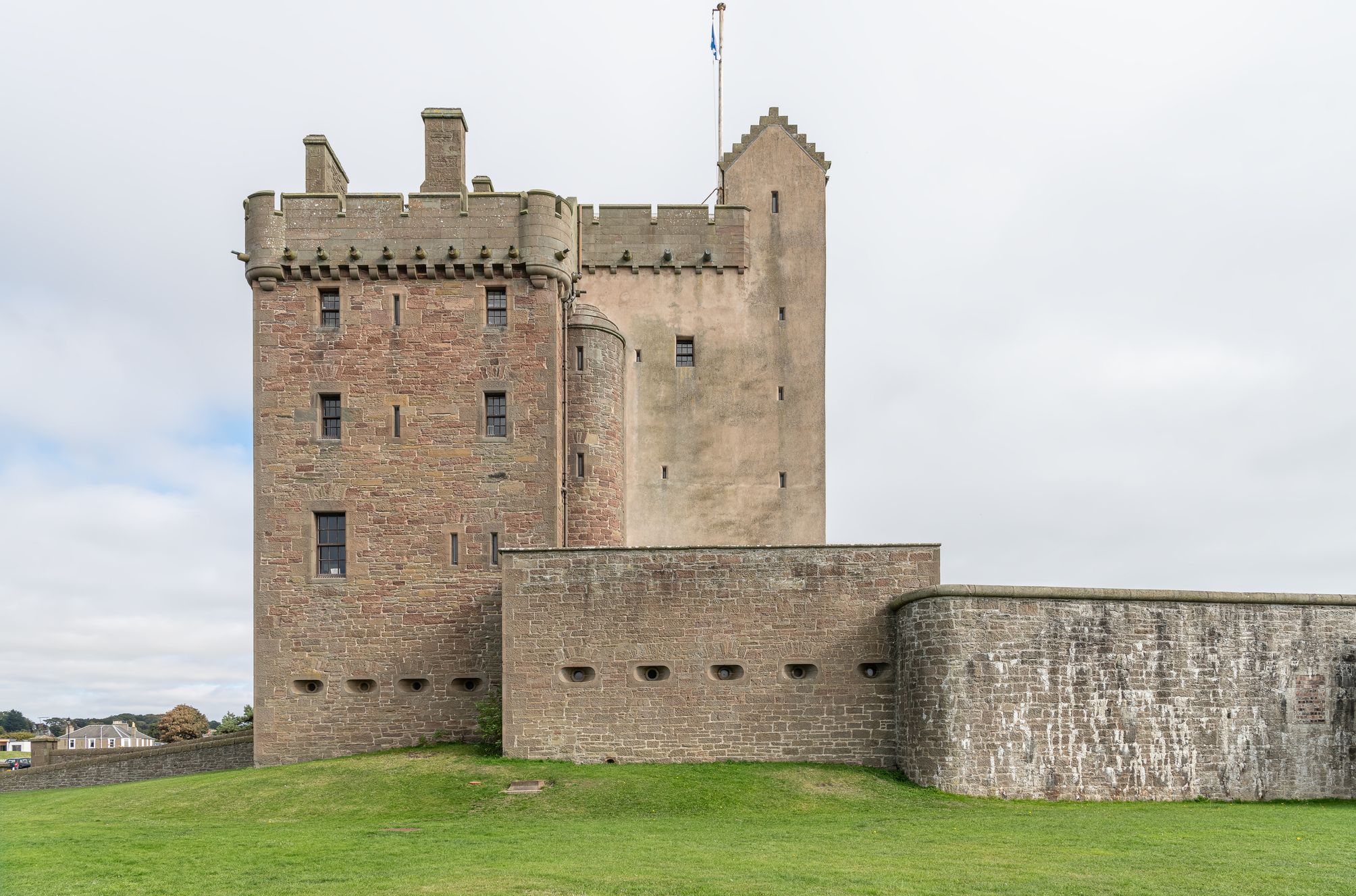 Broughty Castle with crenelated towers, narrow windows, and a flagpole flying a blue and white flag, surrounded by a grassy lawn under an overcast sky.