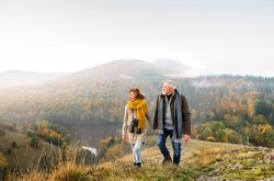 An older couple walking on a grassy hill, dressed warmly with scarves and jackets, enjoying a misty autumn landscape of colorful trees and rolling hills in the background.