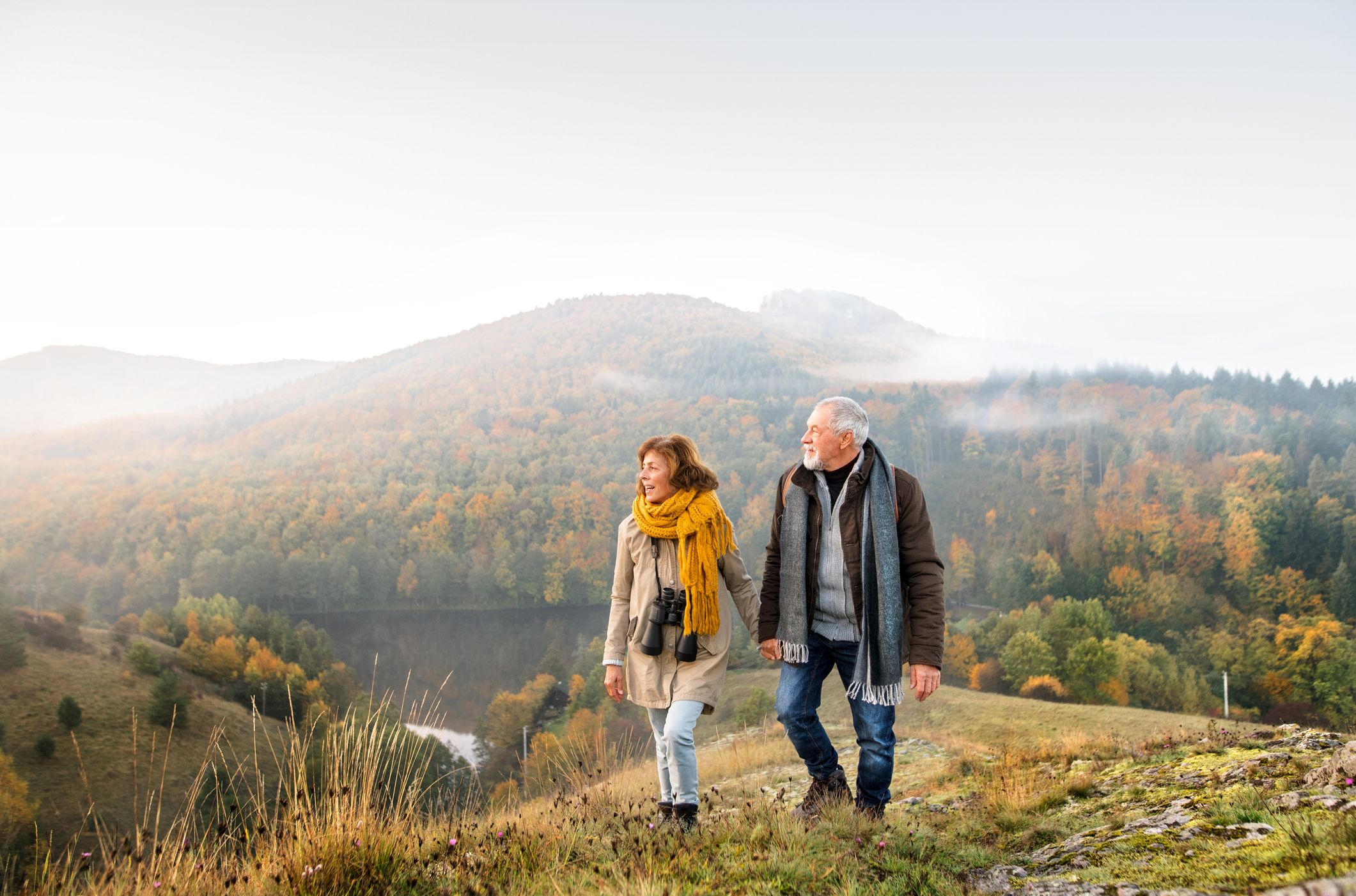 An older couple walking on a grassy hill, dressed warmly with scarves and jackets, enjoying a misty autumn landscape of colorful trees and rolling hills in the background.