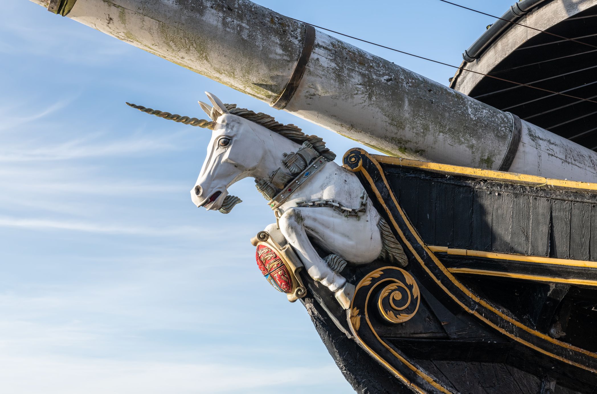 Close-up of a ship's figurehead shaped like a white unicorn with a golden horn, ornate detailing, and a red crest on a black and gold hull, set against a clear blue sky.