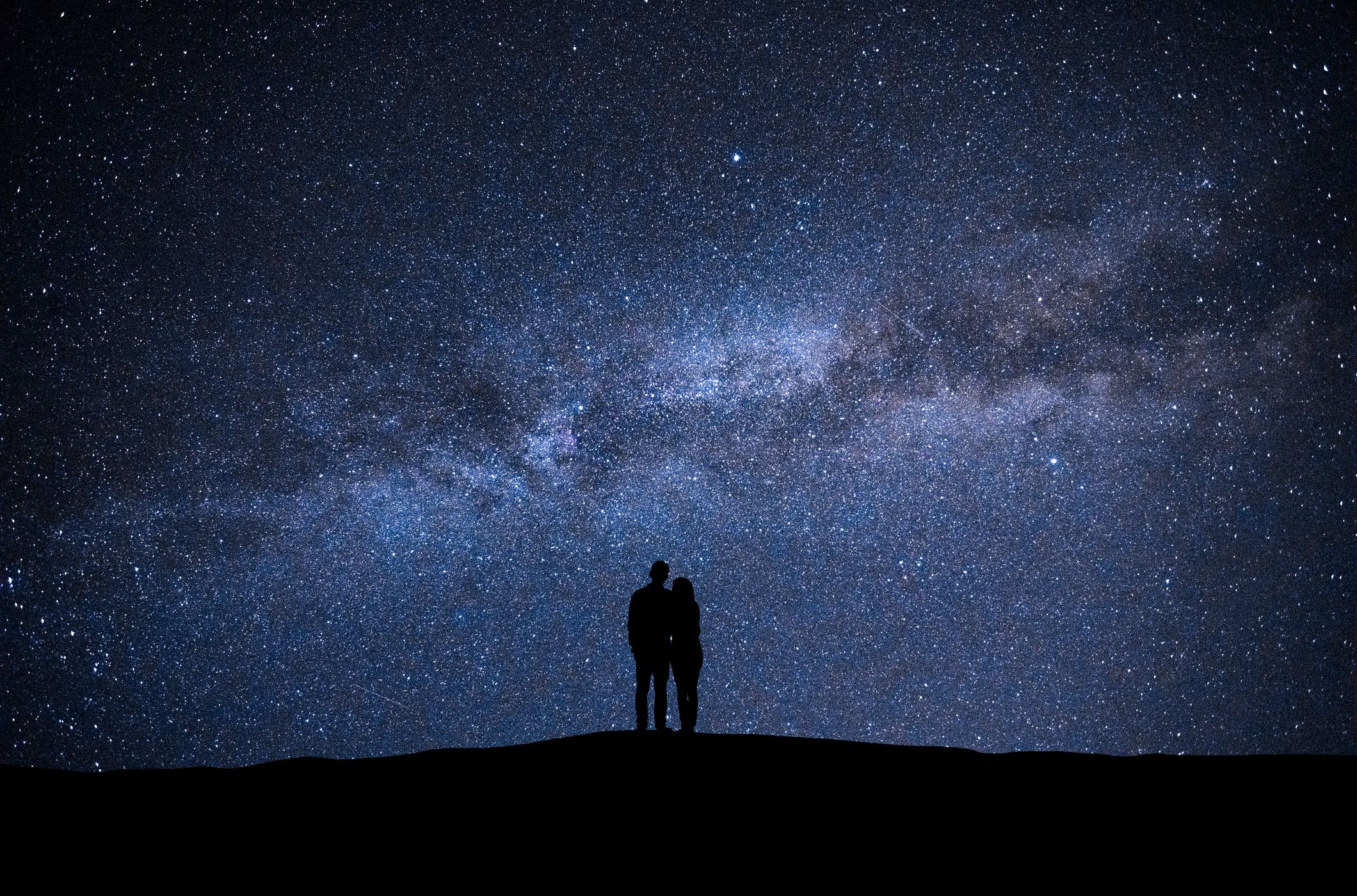 Silhouette of a couple standing on a hill, gazing at a star-filled night sky with the Milky Way stretching across the horizon in a stunning cosmic display.