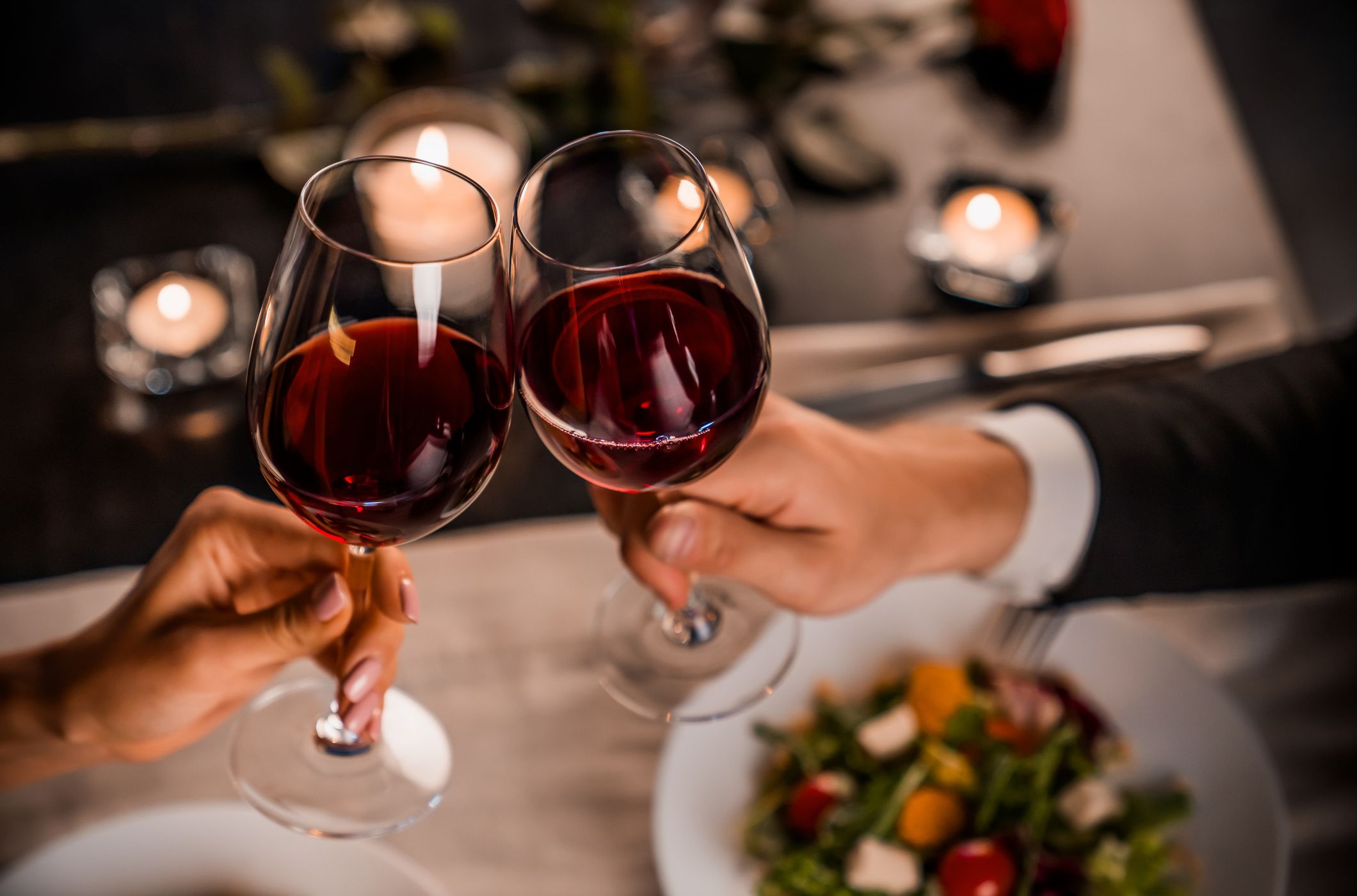 Two hands clinking glasses of red wine over a candlelit dinner table, featuring plates of fresh salad and soft lighting creating a romantic ambiance.