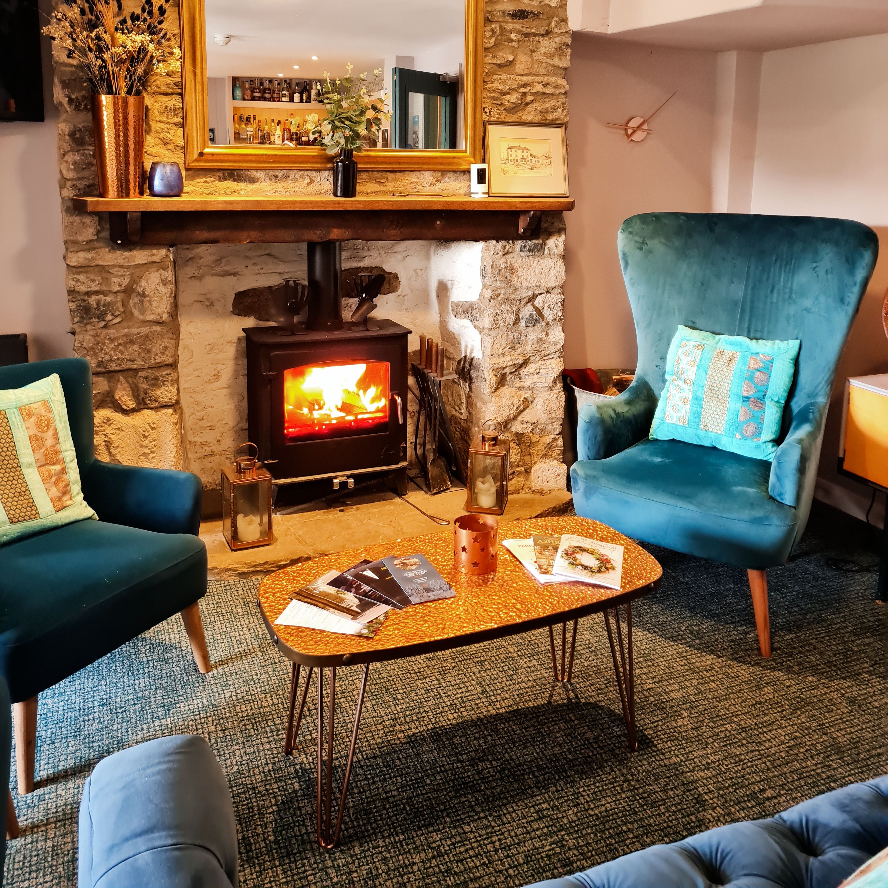 Cozy living room with blue armchairs, a stone fireplace with a wood-burning stove, a large mirror, and a coffee table with magazines.
