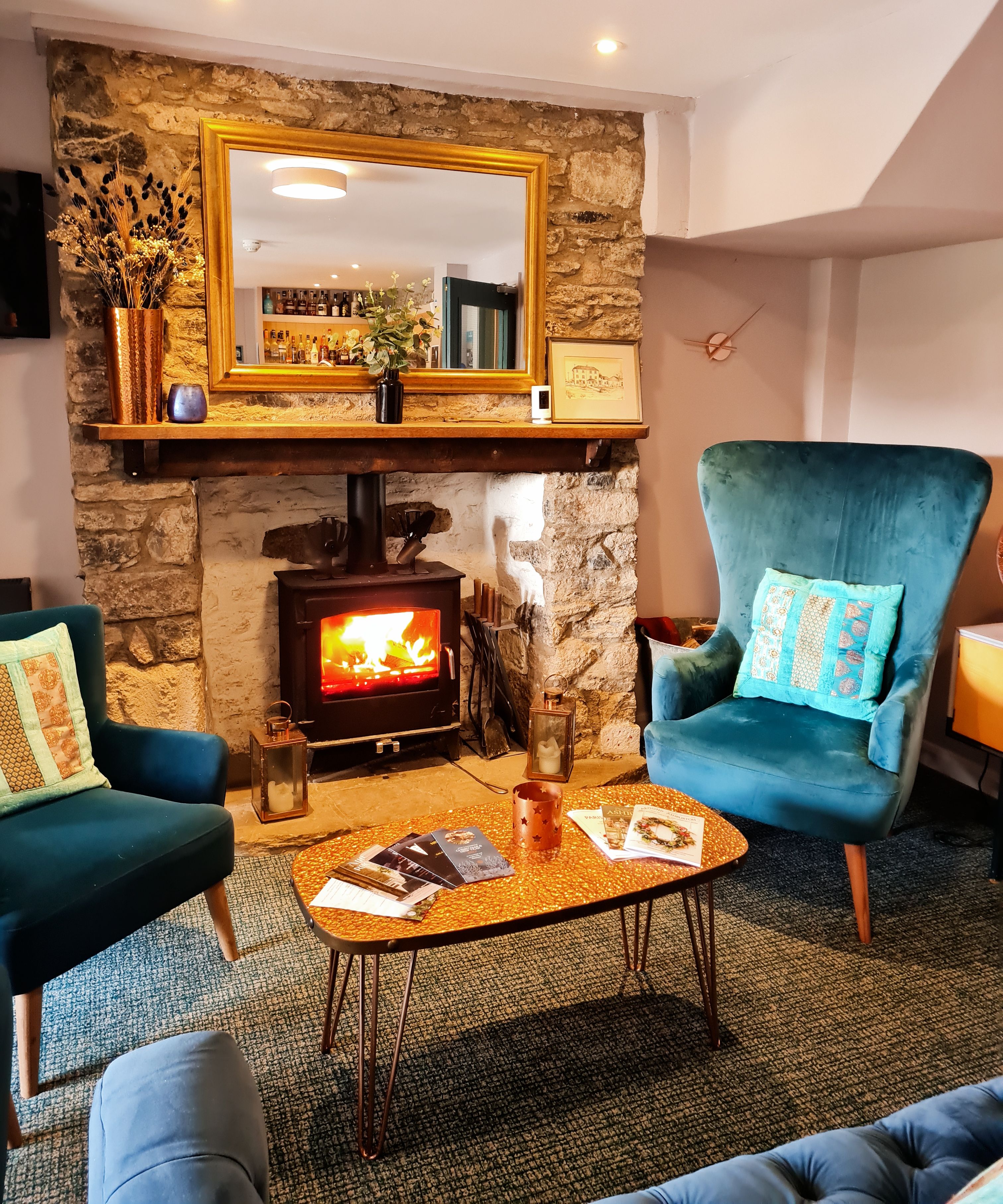 Cozy living room with blue armchairs, a stone fireplace with a wood-burning stove, a large mirror, and a coffee table with magazines.