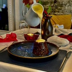 Pouring sauce over a rich dessert pudding on a table set for a festive meal.