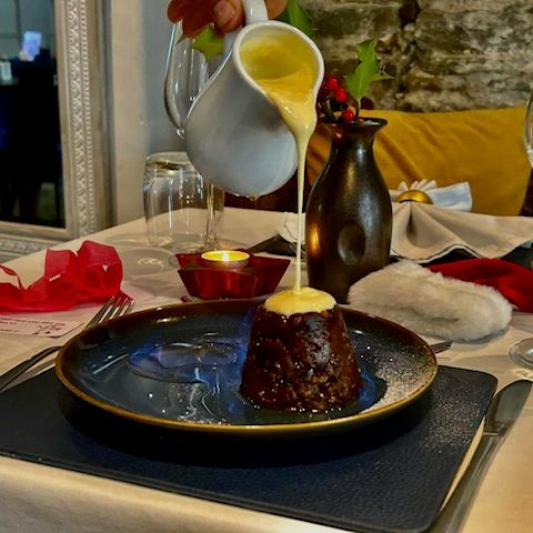 Pouring sauce over a rich dessert pudding on a table set for a festive meal.