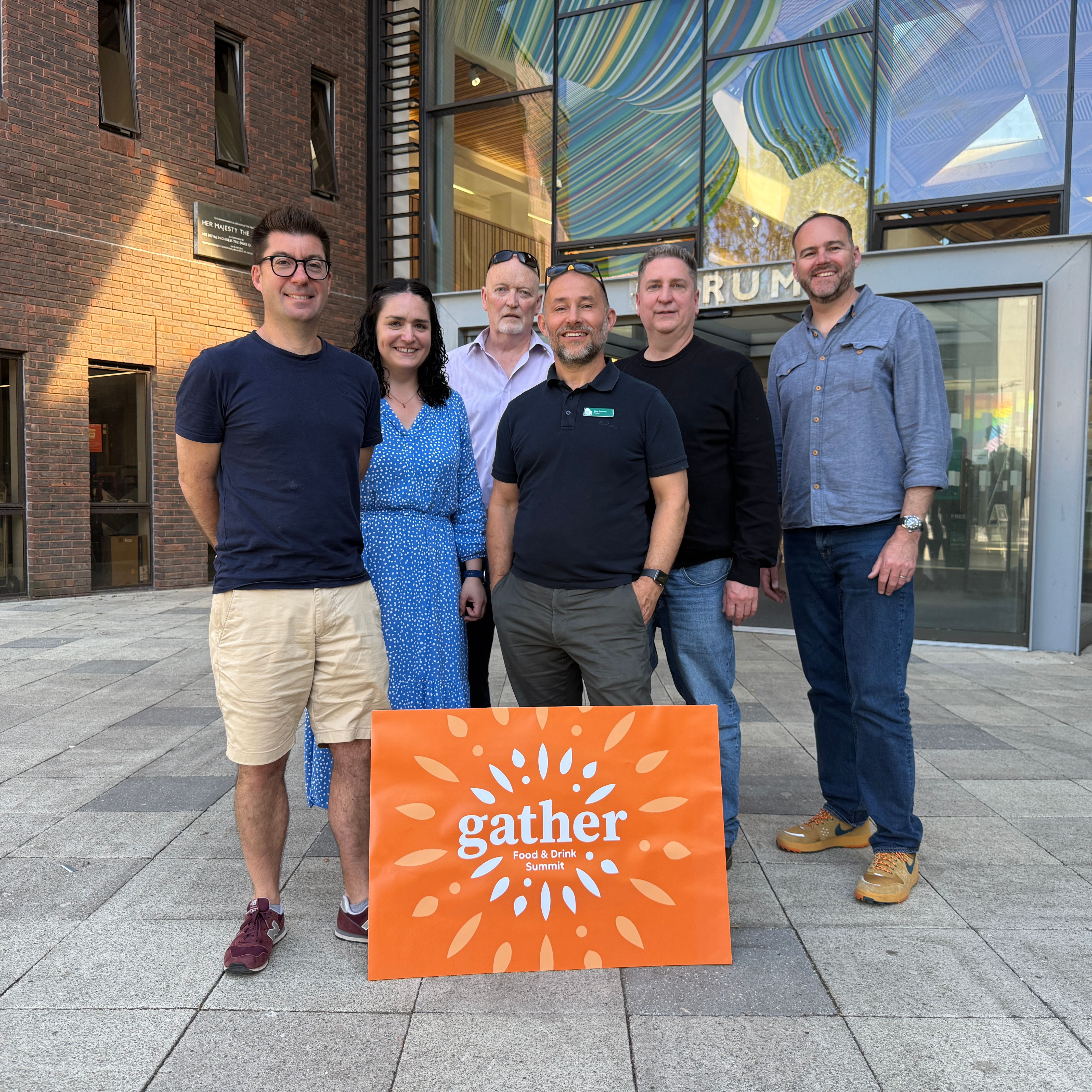 Six people standing outside a modern building, posing with an orange sign that says 'gather Food Drink Summit'.