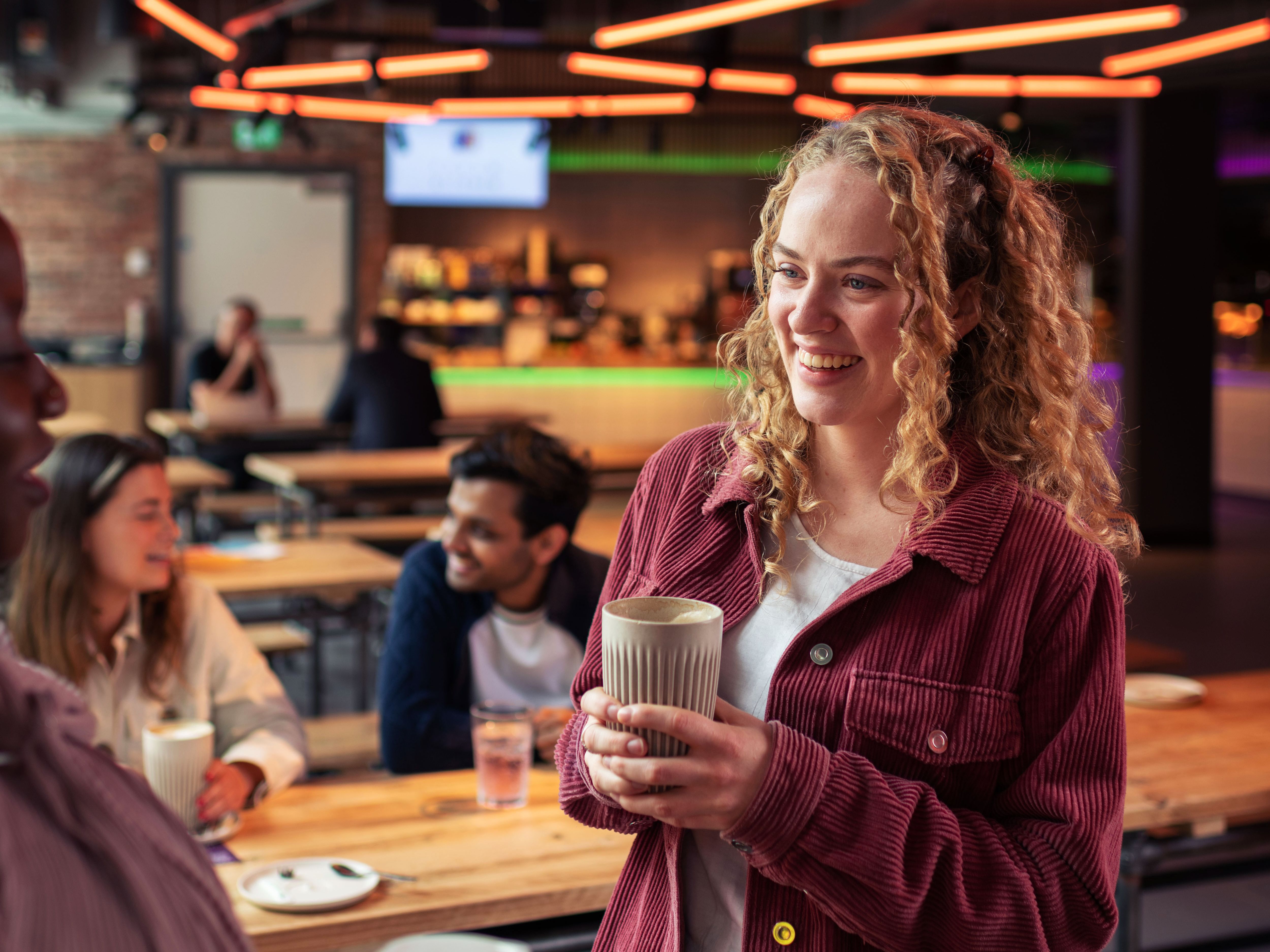 Group of young adults socializing and holding drinks in a modern bar or café setting with neon lights overhead.