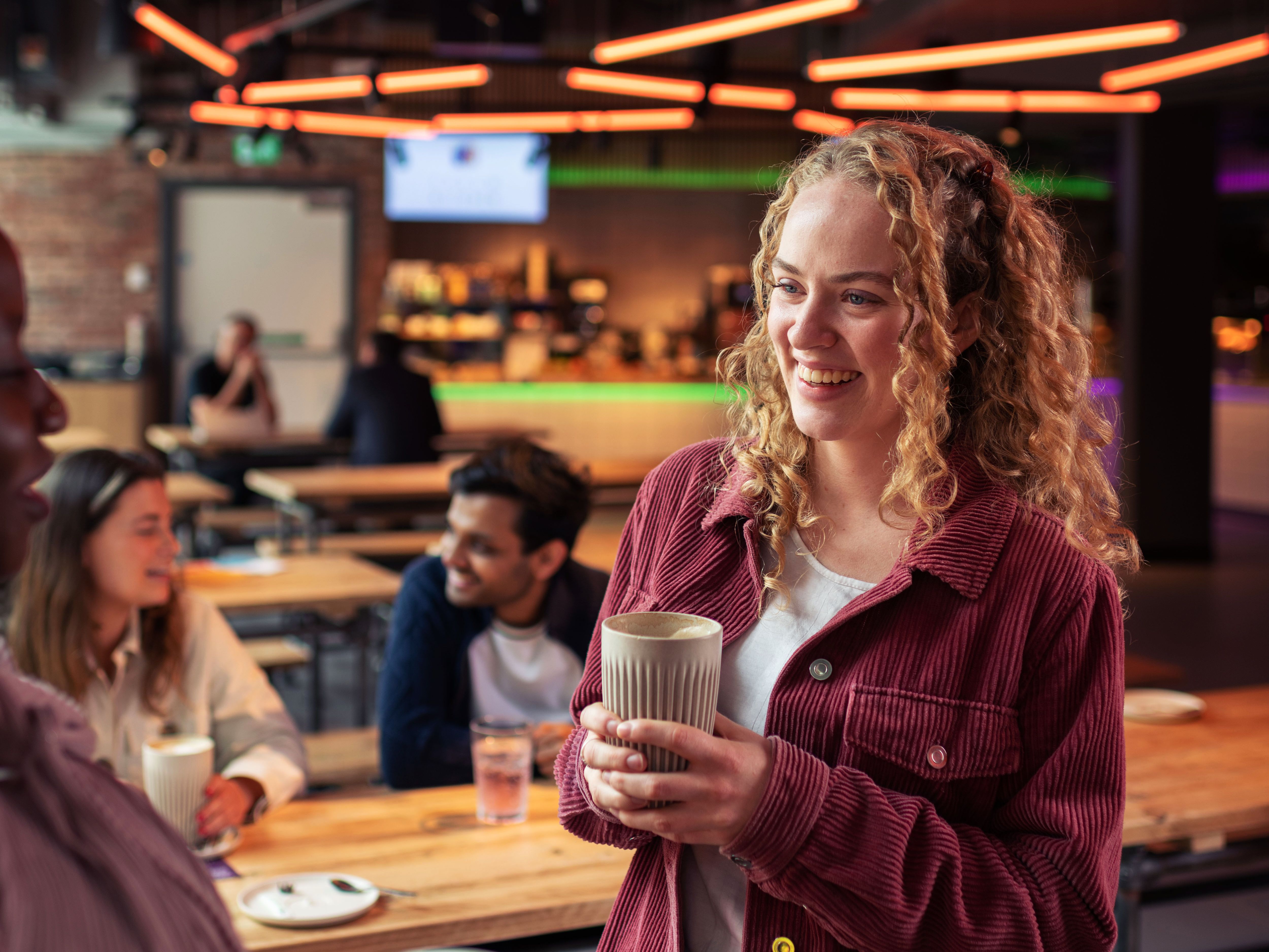 Group of young adults socializing and holding drinks in a modern bar or café setting with neon lights overhead.