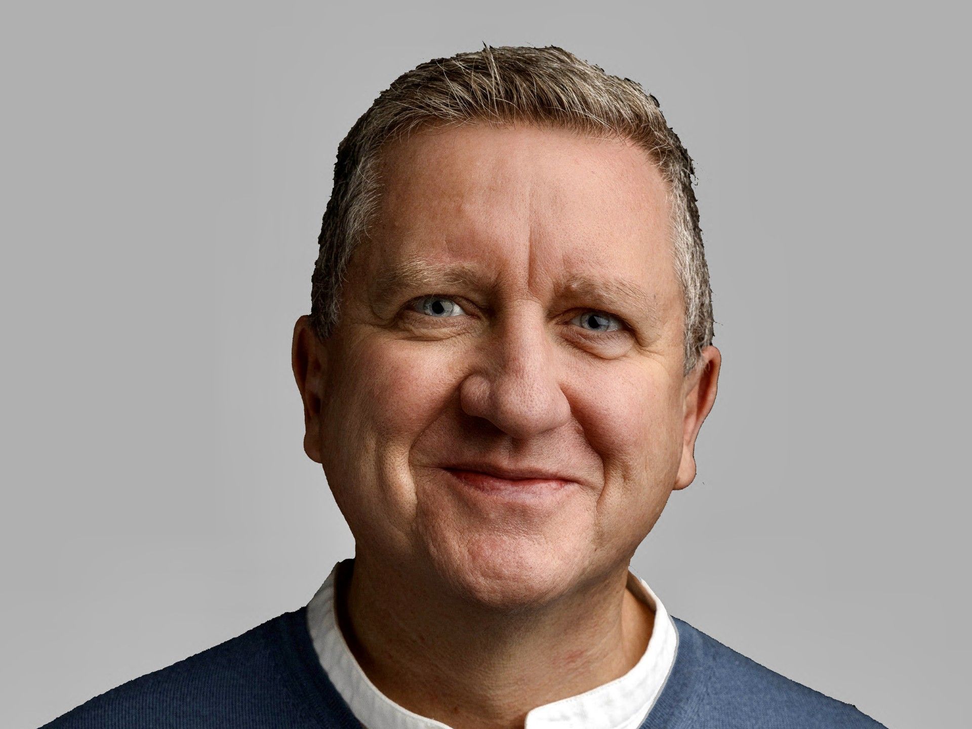 Smiling middle-aged man in a blue sweater and white shirt against a neutral gray background.