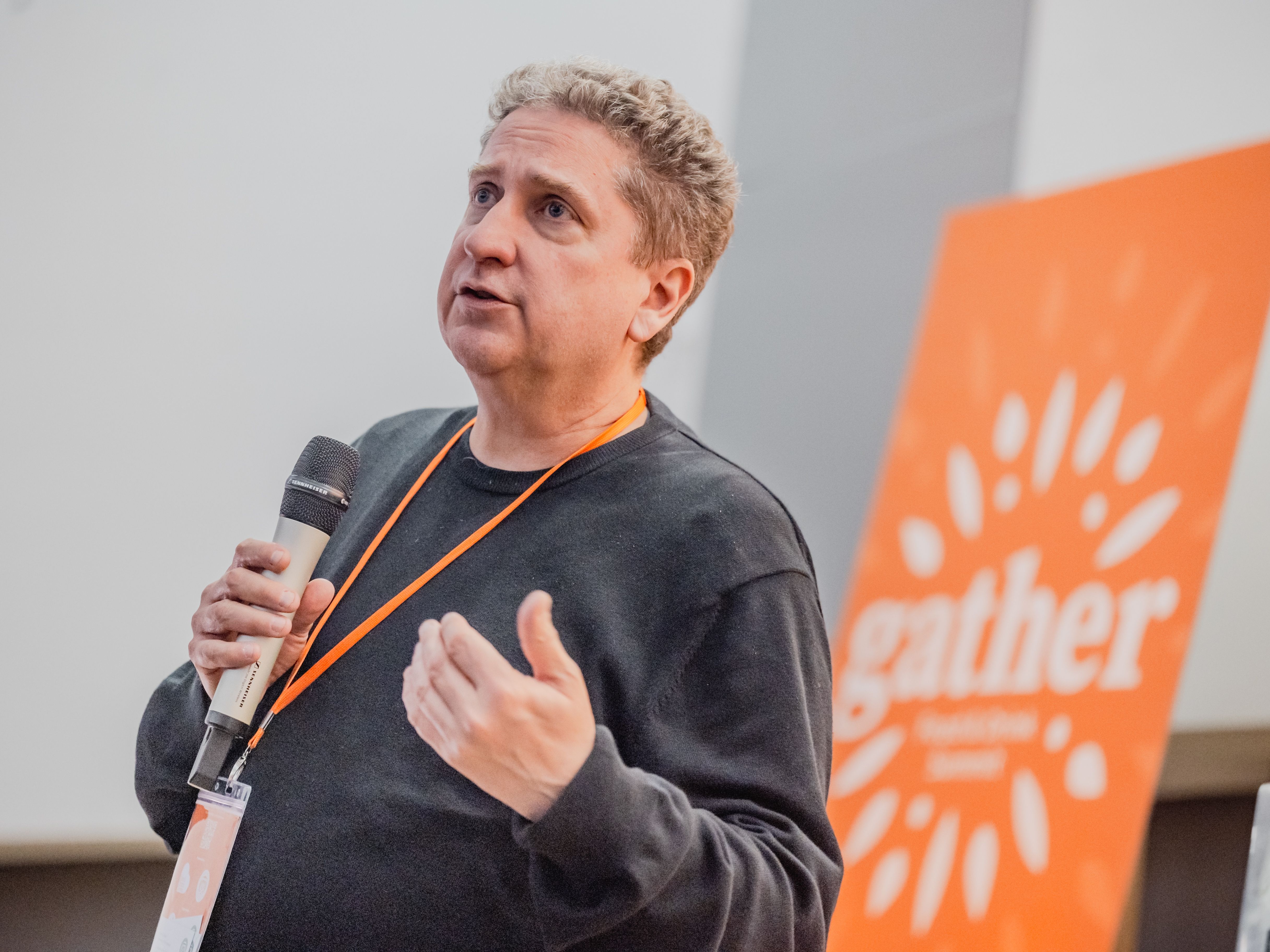 A person holding a microphone and speaking at an event, with an orange 'gather' sign in the background.