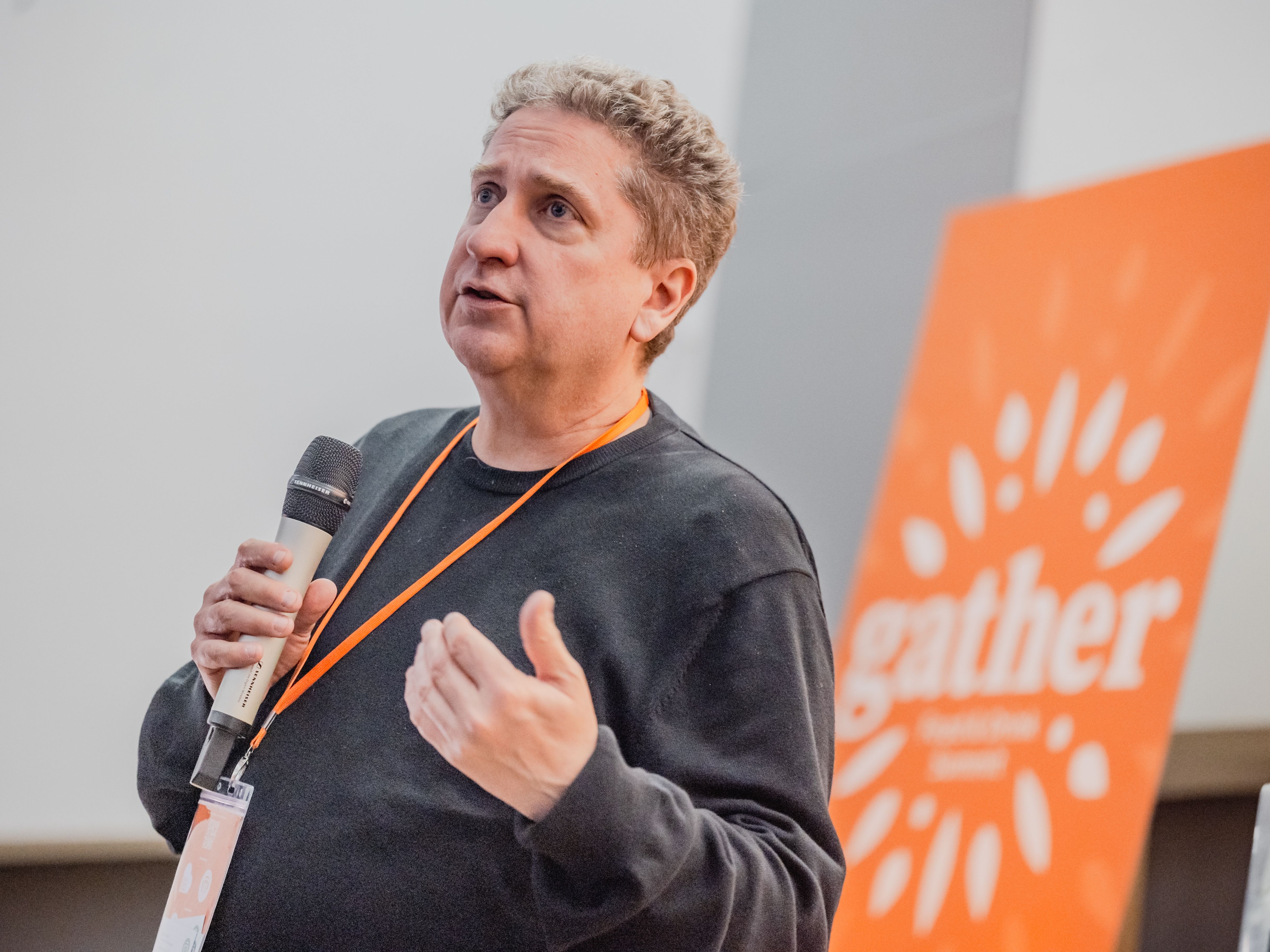 A person holding a microphone and speaking at an event, with an orange 'gather' sign in the background.