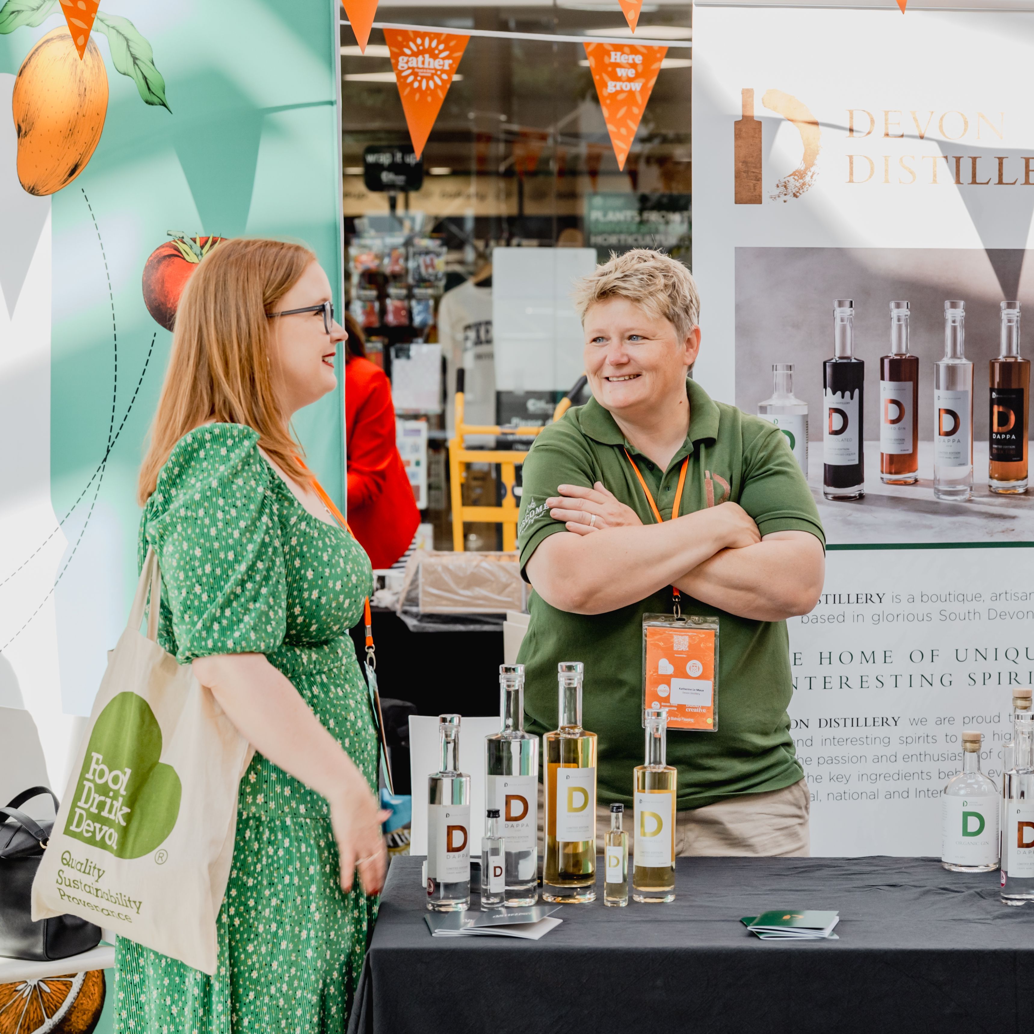 Two people talking at a distillery booth at a market or event, with a display of bottles and a Devon Distillery banner.