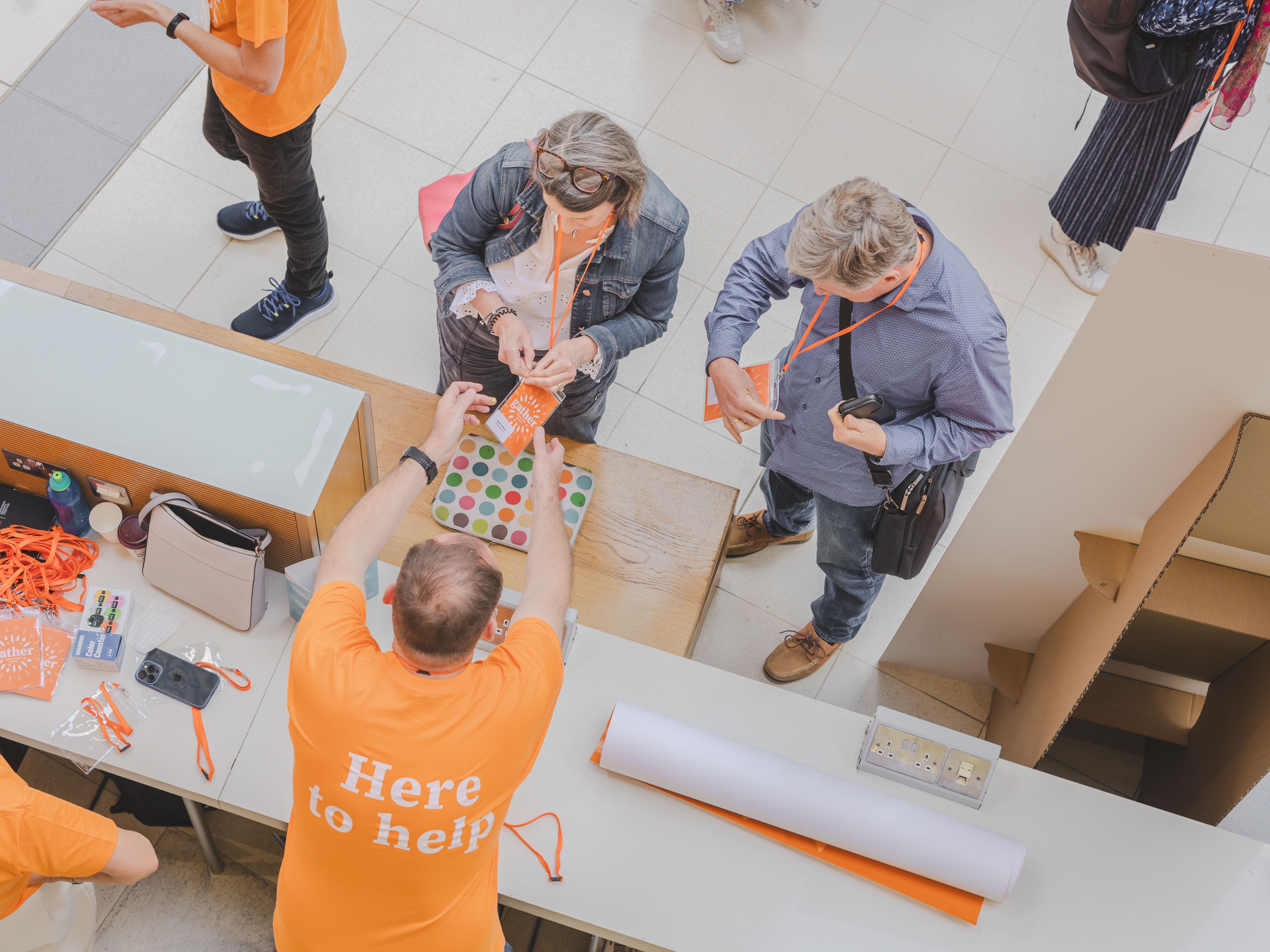 Event registration desk with volunteers in orange 'Here to help' shirts assisting attendees.