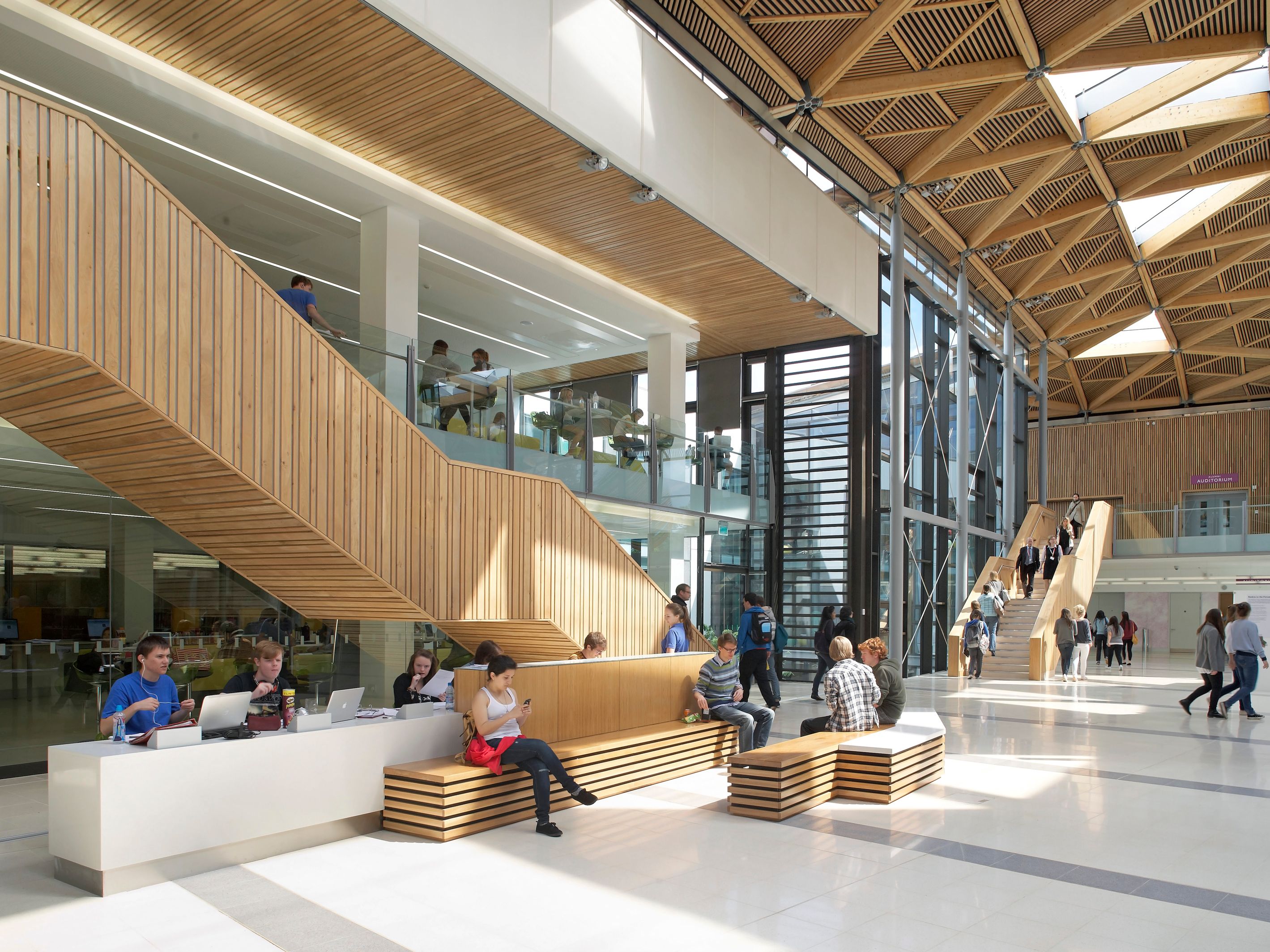 Modern university building interior with students studying and socializing, wooden accents and geometric ceiling design.