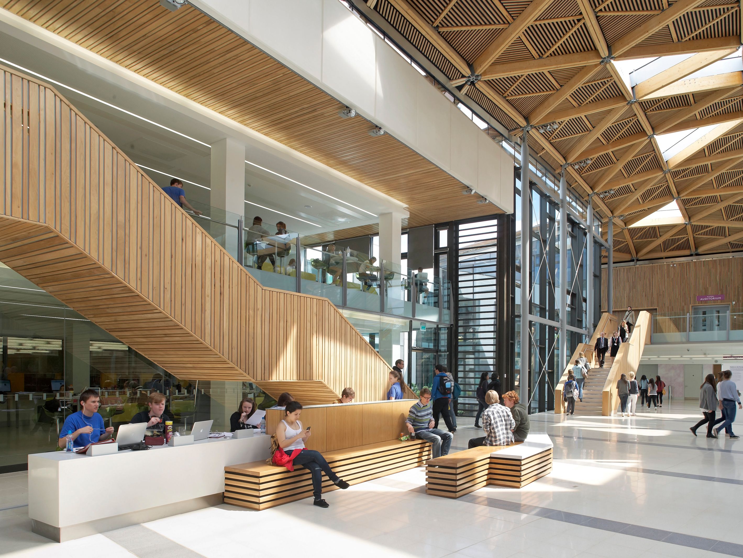 Modern university building interior with students studying and socializing, wooden accents and geometric ceiling design.