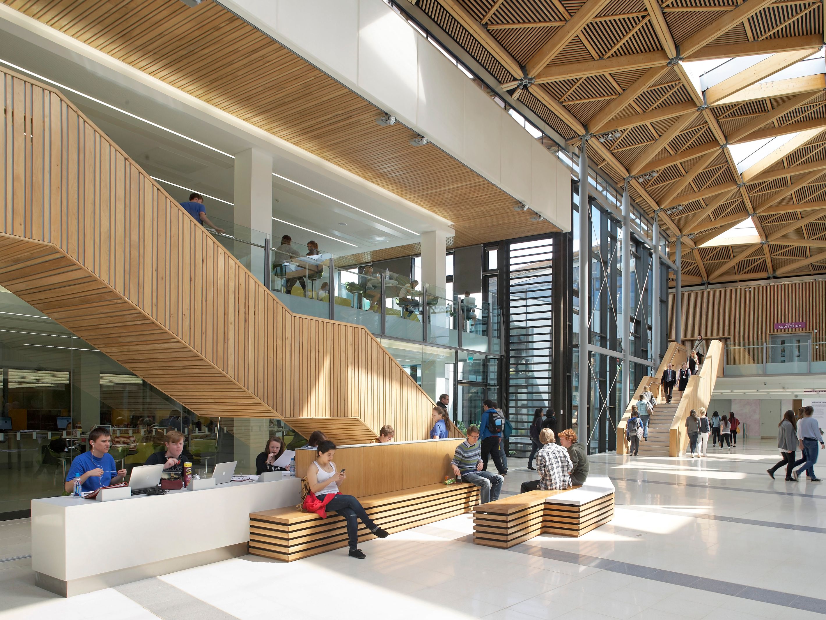 Modern university building interior with students studying and socializing, wooden accents and geometric ceiling design.