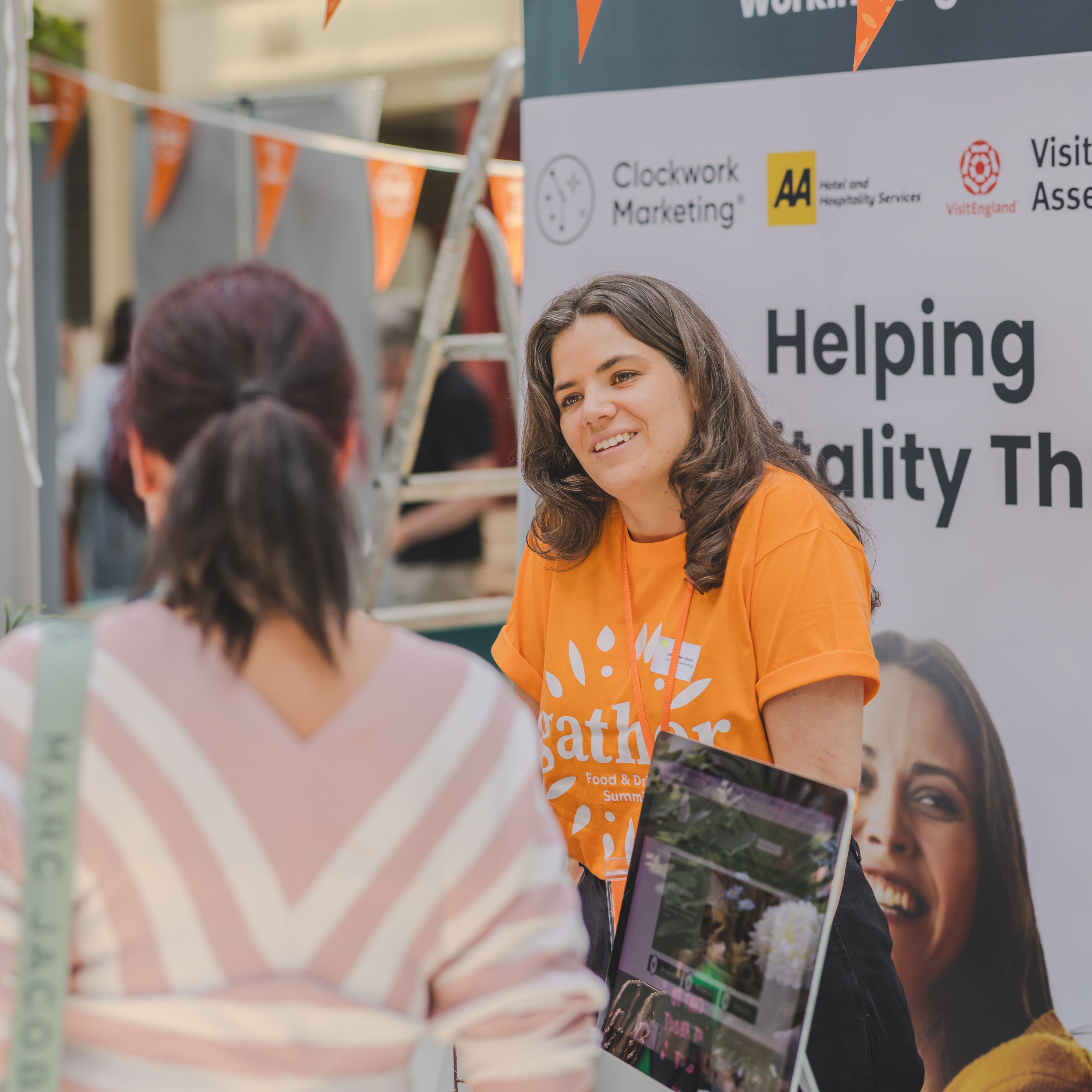 Woman in an orange t-shirt talking to another person at a hospitality event booth.
