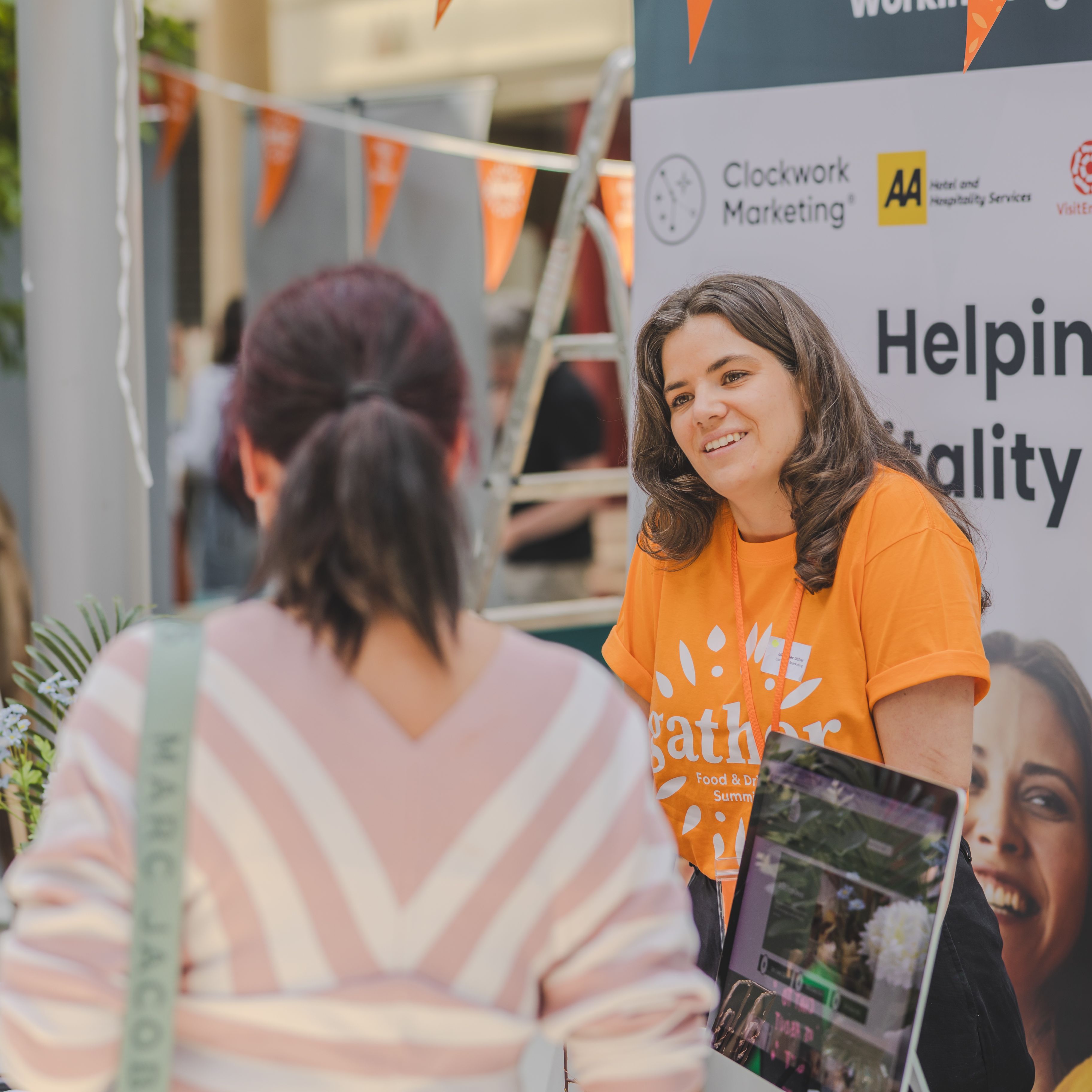 Woman in an orange t-shirt talking to another person at a hospitality event booth.