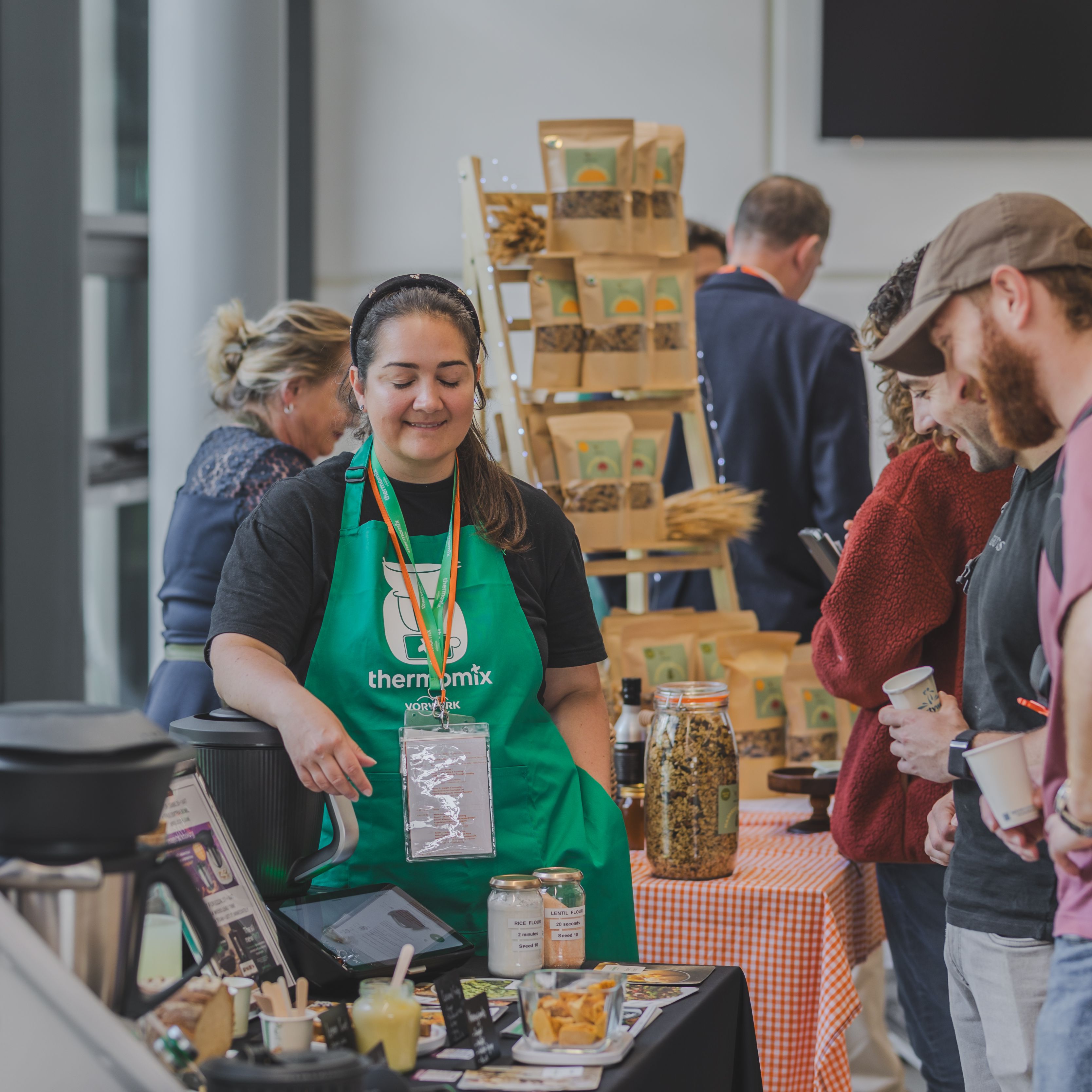Person wearing a green apron at a market or food fair, presenting products to attendees.