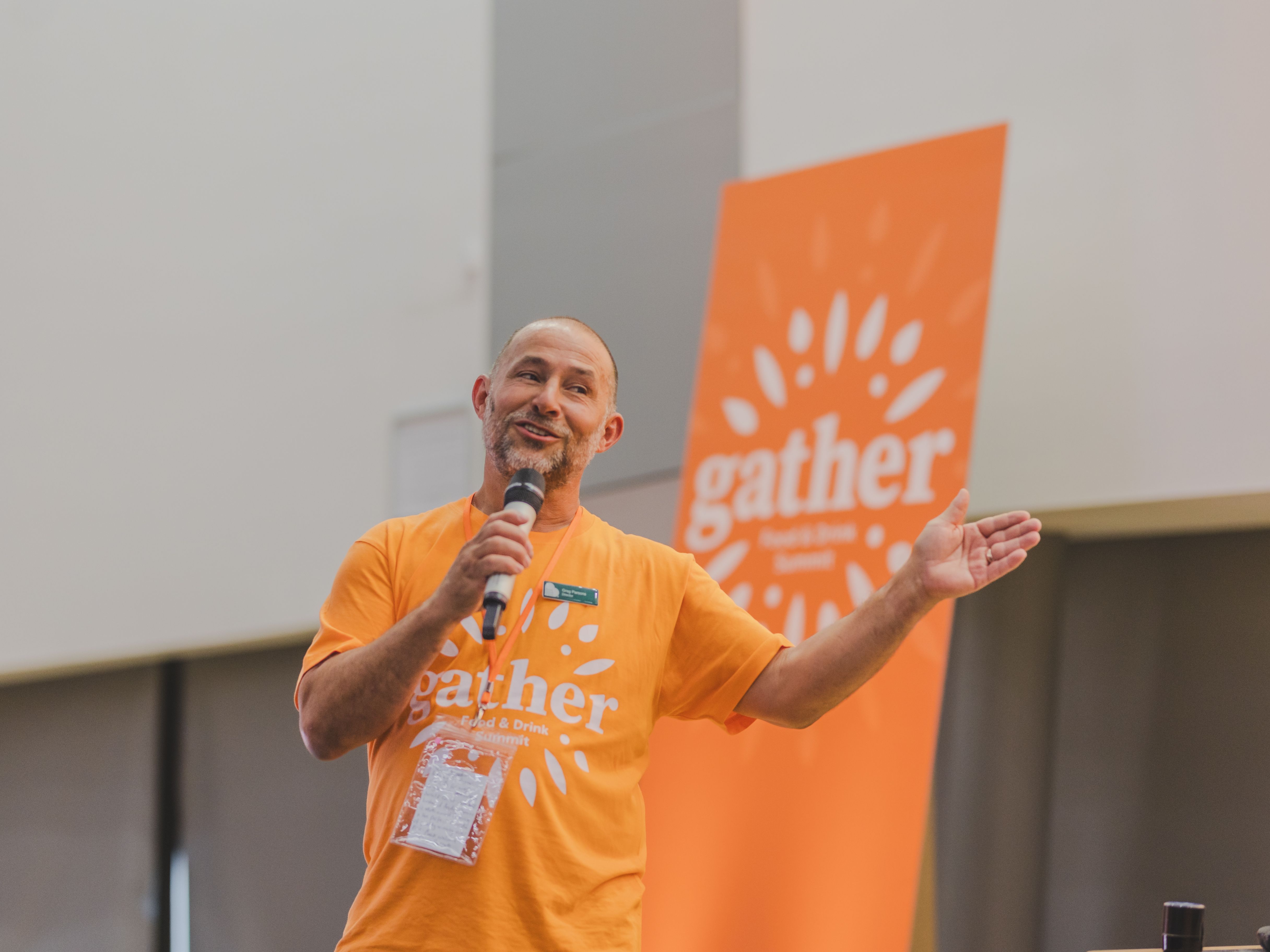 Man speaking into a microphone wearing an orange 'gather' shirt at an event, with a matching orange 'gather' banner in the background.