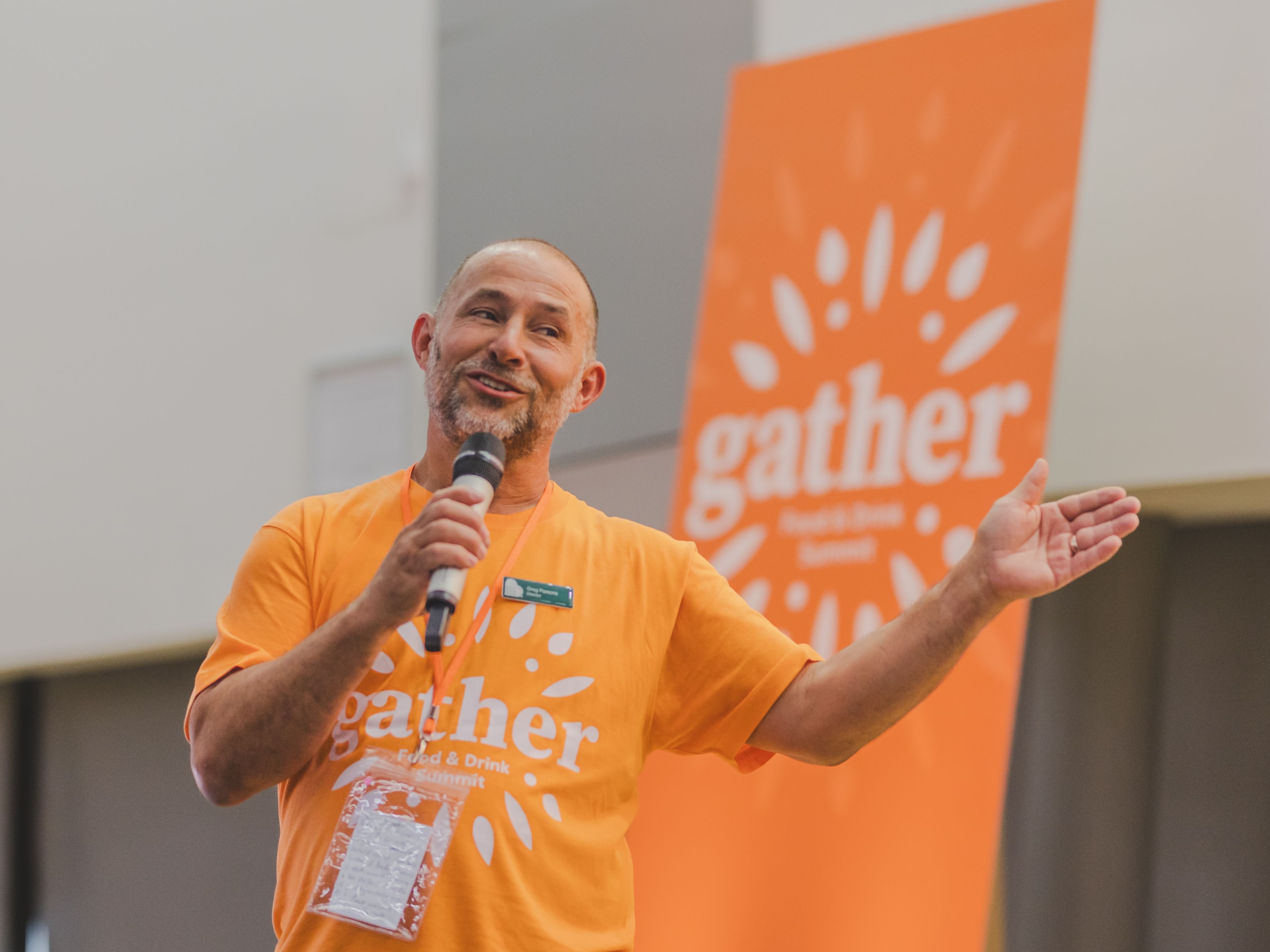 Man speaking into a microphone wearing an orange 'gather' shirt at an event, with a matching orange 'gather' banner in the background.