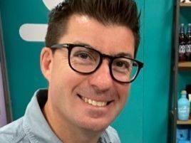 Smiling man with dark hair and glasses standing in front of a teal background.