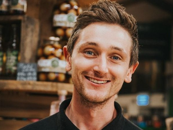 Smiling young man with short brown hair standing indoors, with shelves of jars in the background.