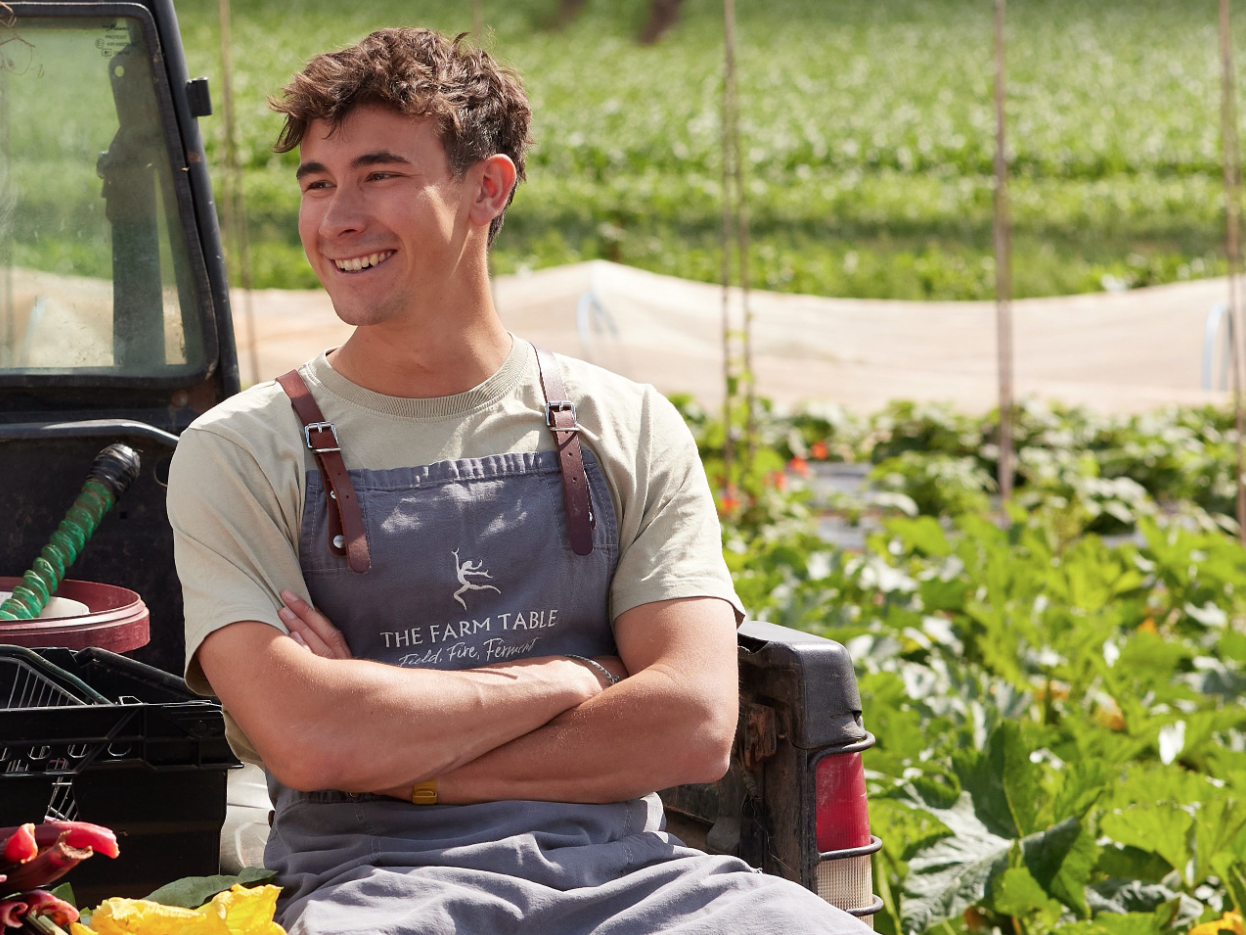 Smiling person wearing an apron that says 'The Farm Table' sitting in a garden or farm setting.