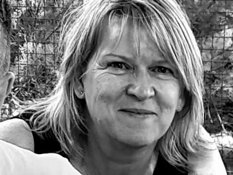 Smiling woman with light hair in a black top posing outdoors with two people partially visible beside her