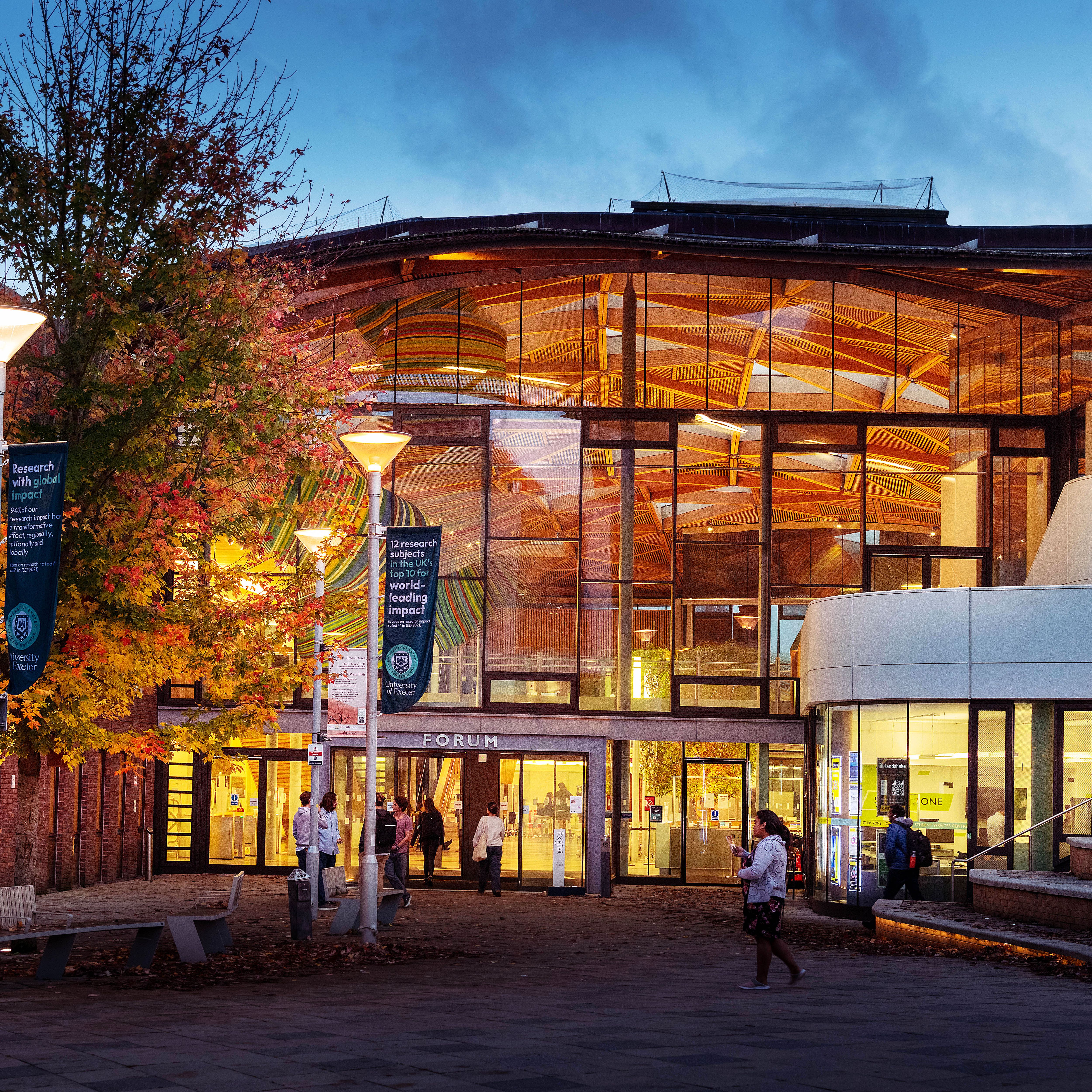 Modern glass building with illuminated interior, people walking outside, trees with autumn leaves.