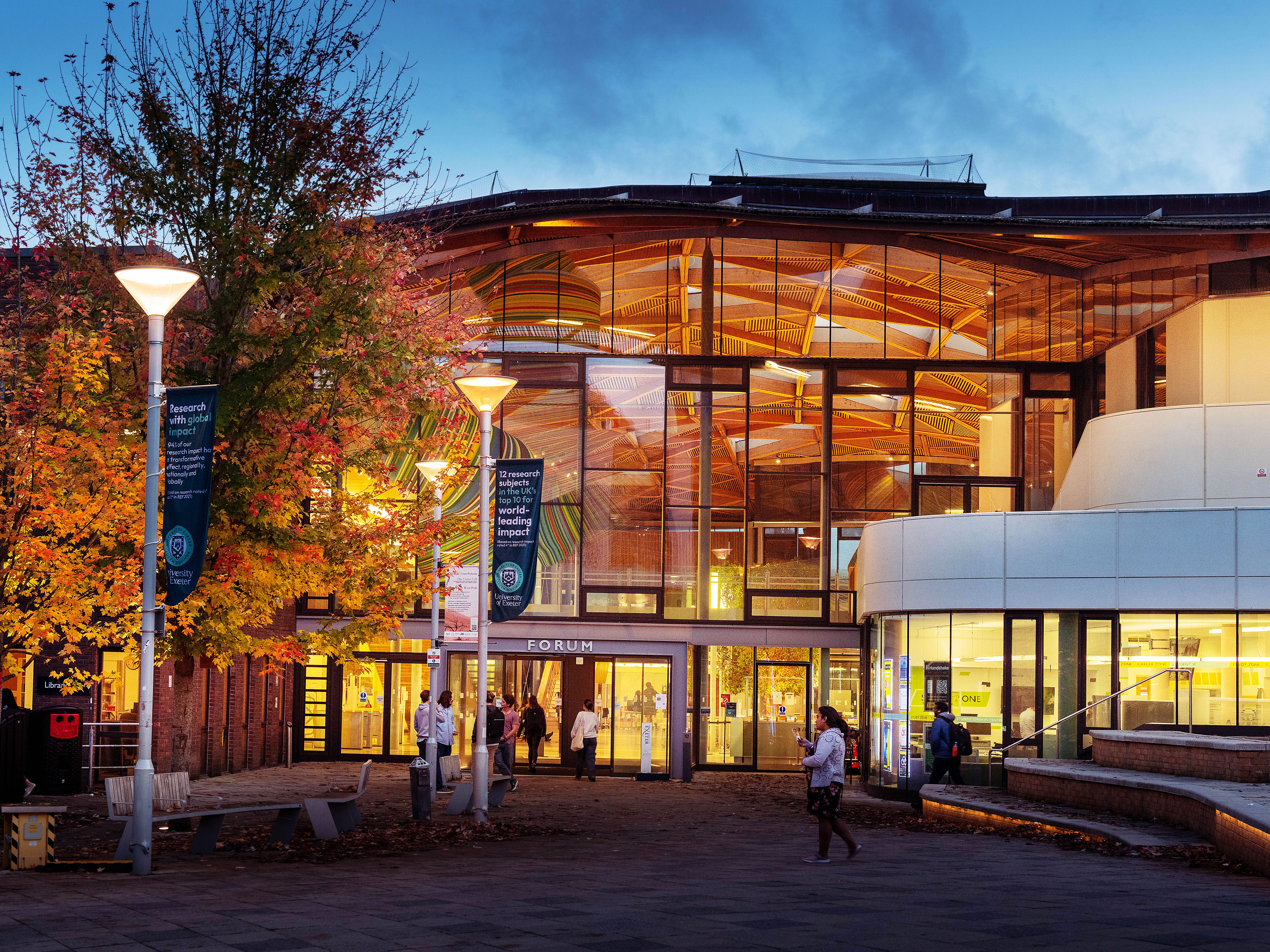 Modern glass building with illuminated interior, people walking outside, trees with autumn leaves.