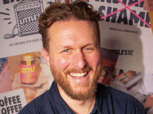 Smiling man with beard and curly hair standing in front of a wall covered with posters and images.