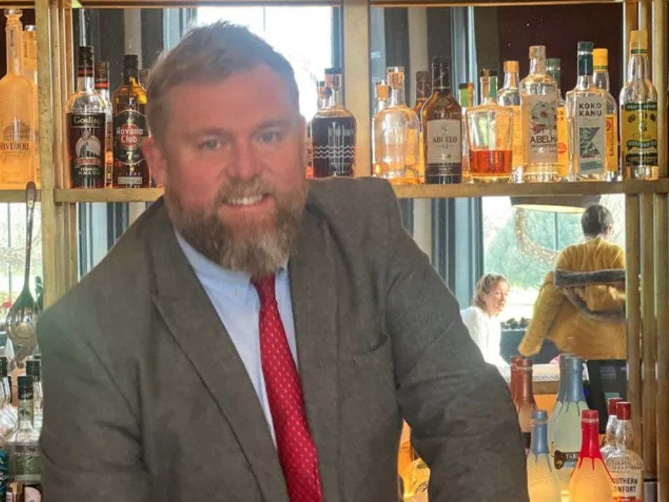 Man in a suit and red tie standing behind a bar with various bottles of alcohol on shelves.