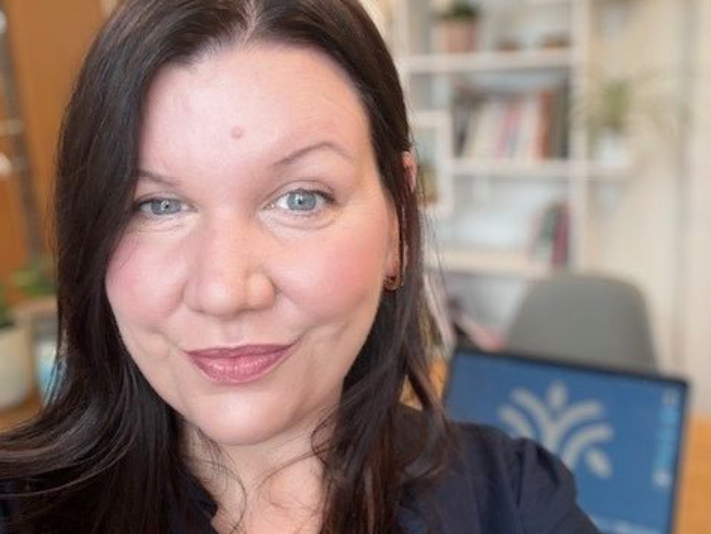 Woman with dark hair smiling in an office setting with a blurred laptop and bookshelf in the background